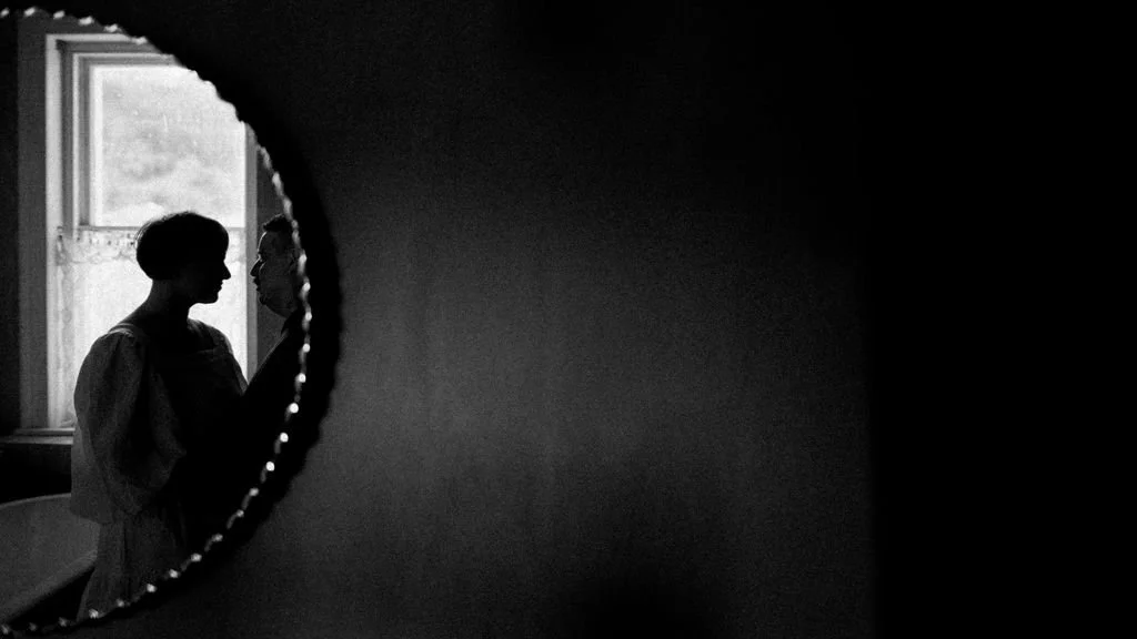 Silhouette of two women facing each other in front of a window, seen through a round hole in a dark wall.