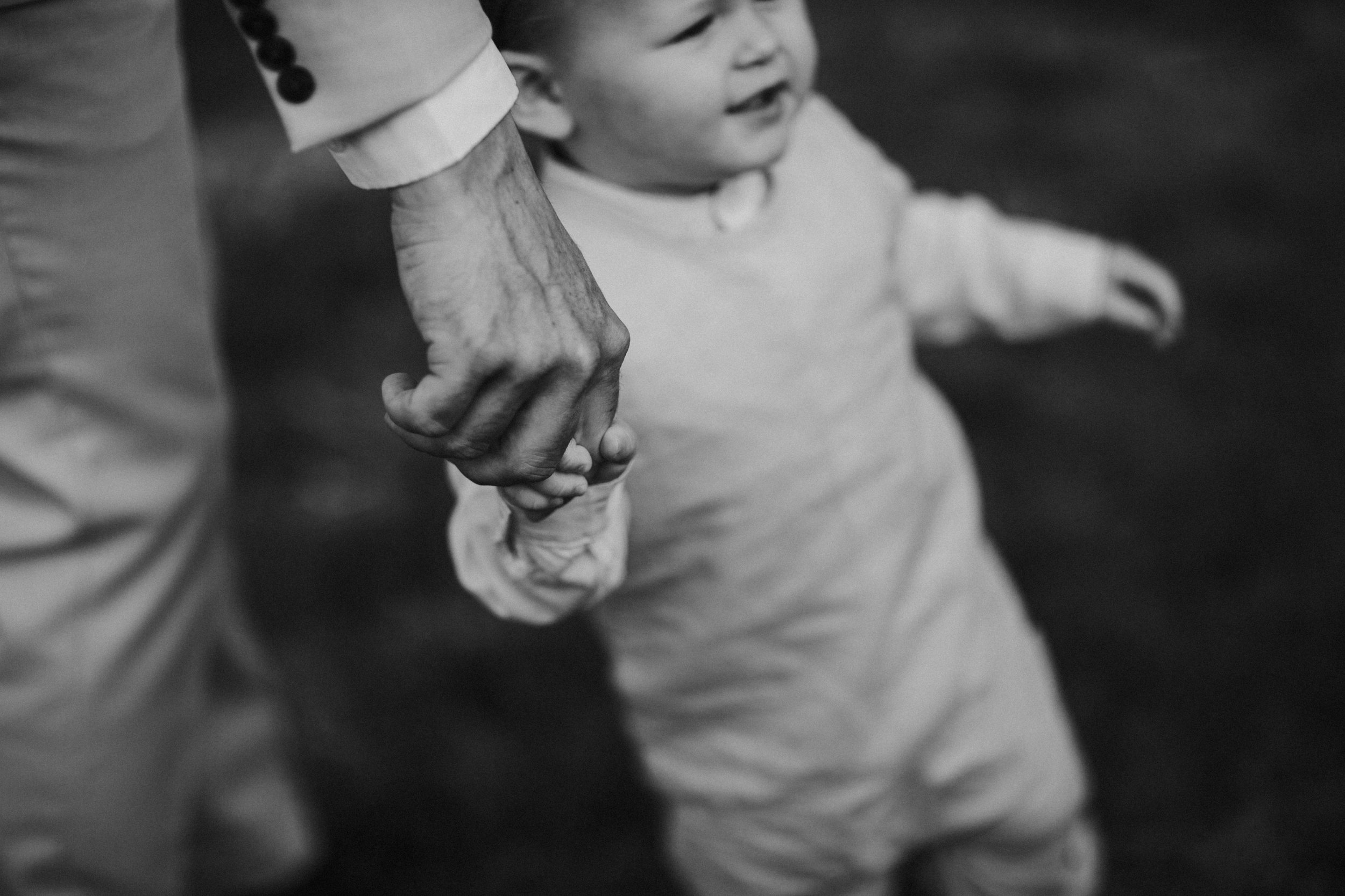 A young child is holding an adult's hand, with the adult's hand covering the child's hand. The child's face is slightly smiling and looking to the side.
