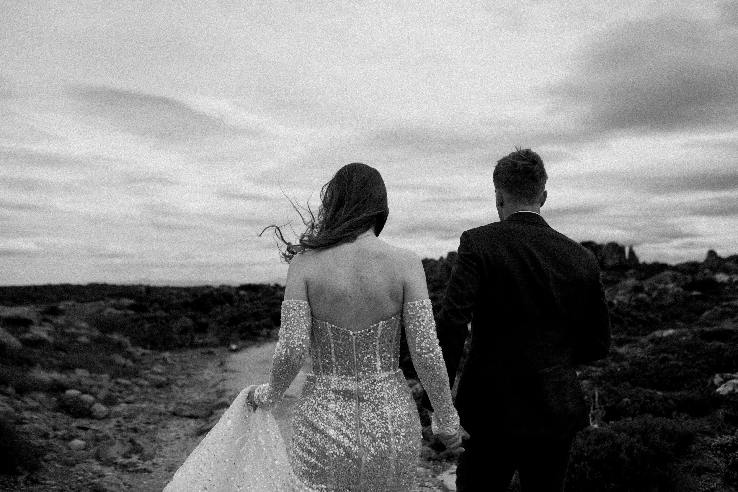 A black and white photo of a bride and groom walking hand in hand on a rocky path, with the bride wearing a sparkling off-shoulder gown and the groom in a dark suit, against a cloudy sky.