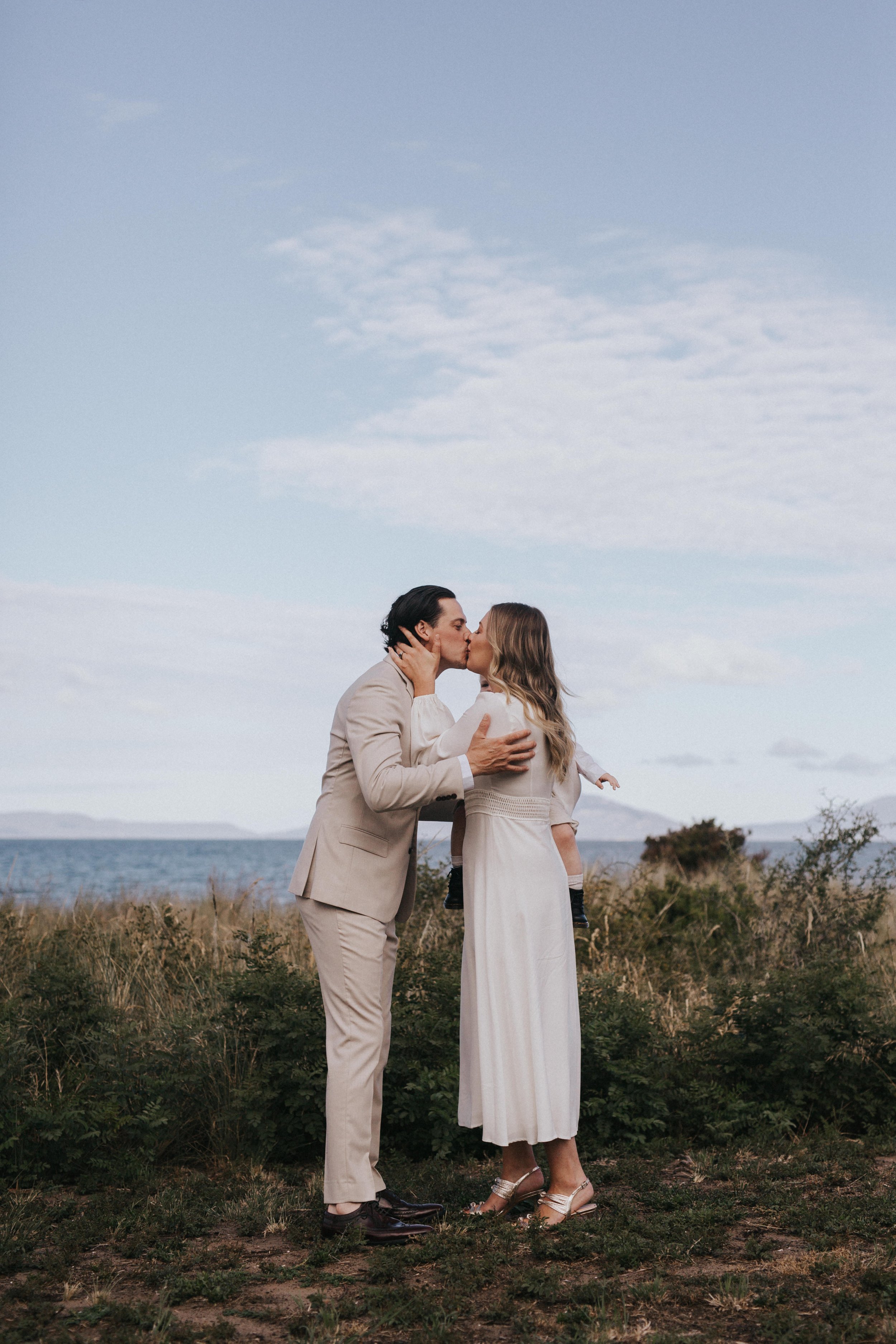 A couple kissing outdoors near water with a person holding a child, under a cloudy sky.