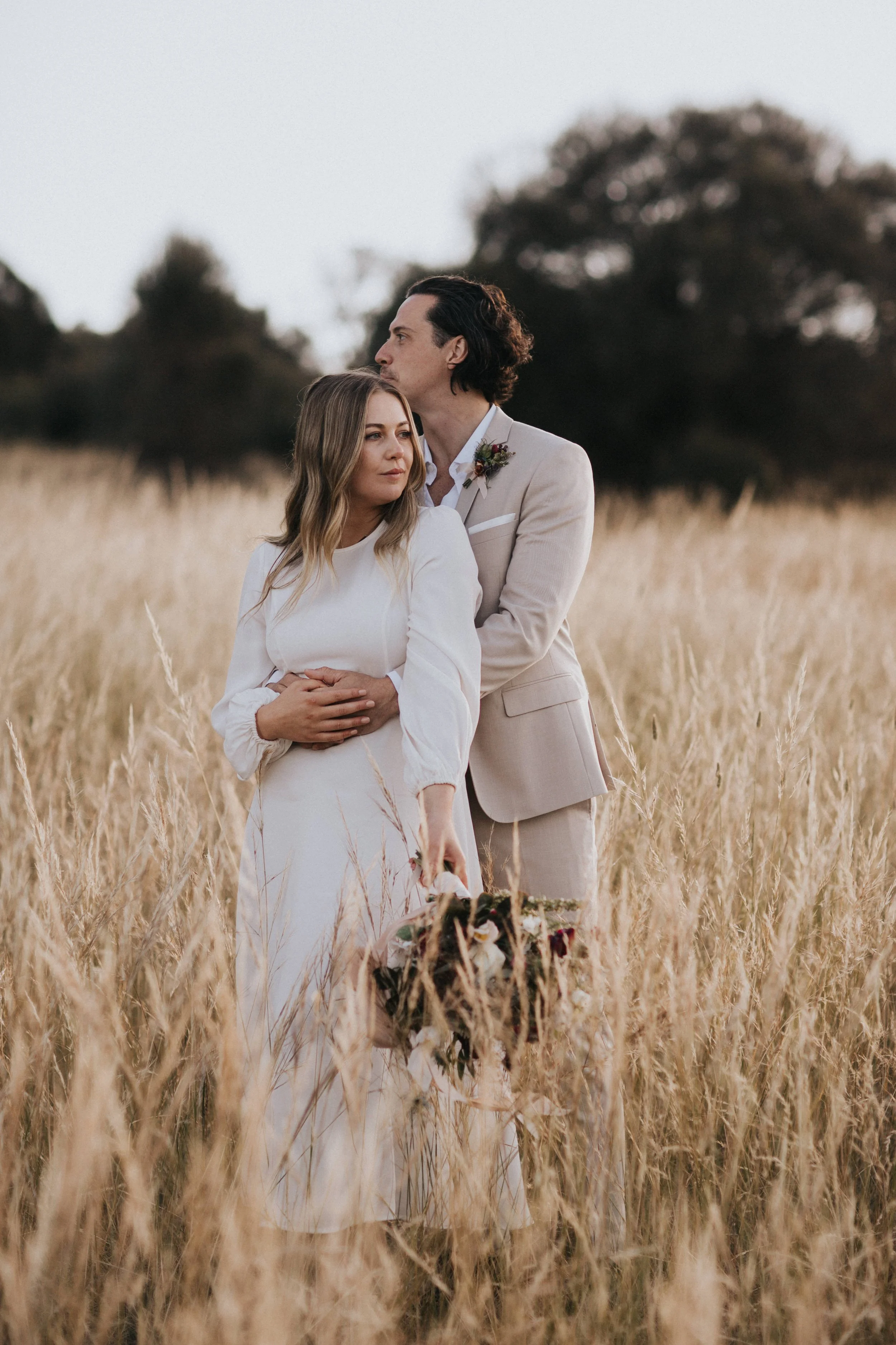 A bride and groom standing in a field of tall, golden grass, with the groom embracing the bride from behind. The bride is holding a bouquet, and both are dressed in wedding attire. The background features trees under an overcast sky.