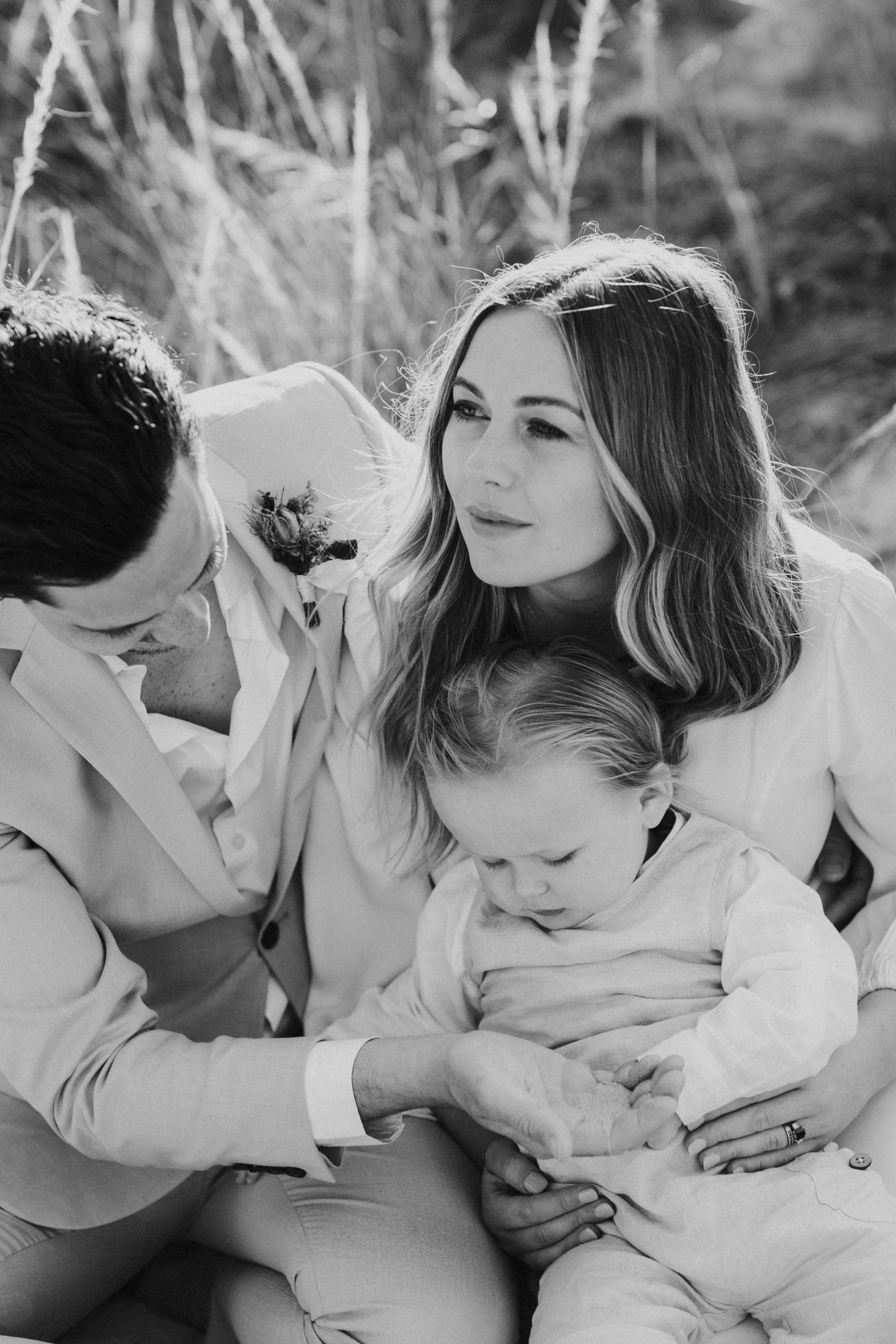 A black-and-white photo of a family outdoors. A man and a woman sit close with a young boy between them. The woman looks at the man, and the boy is focused on something in his hands, while the man reaches out to touch the boy's hand.