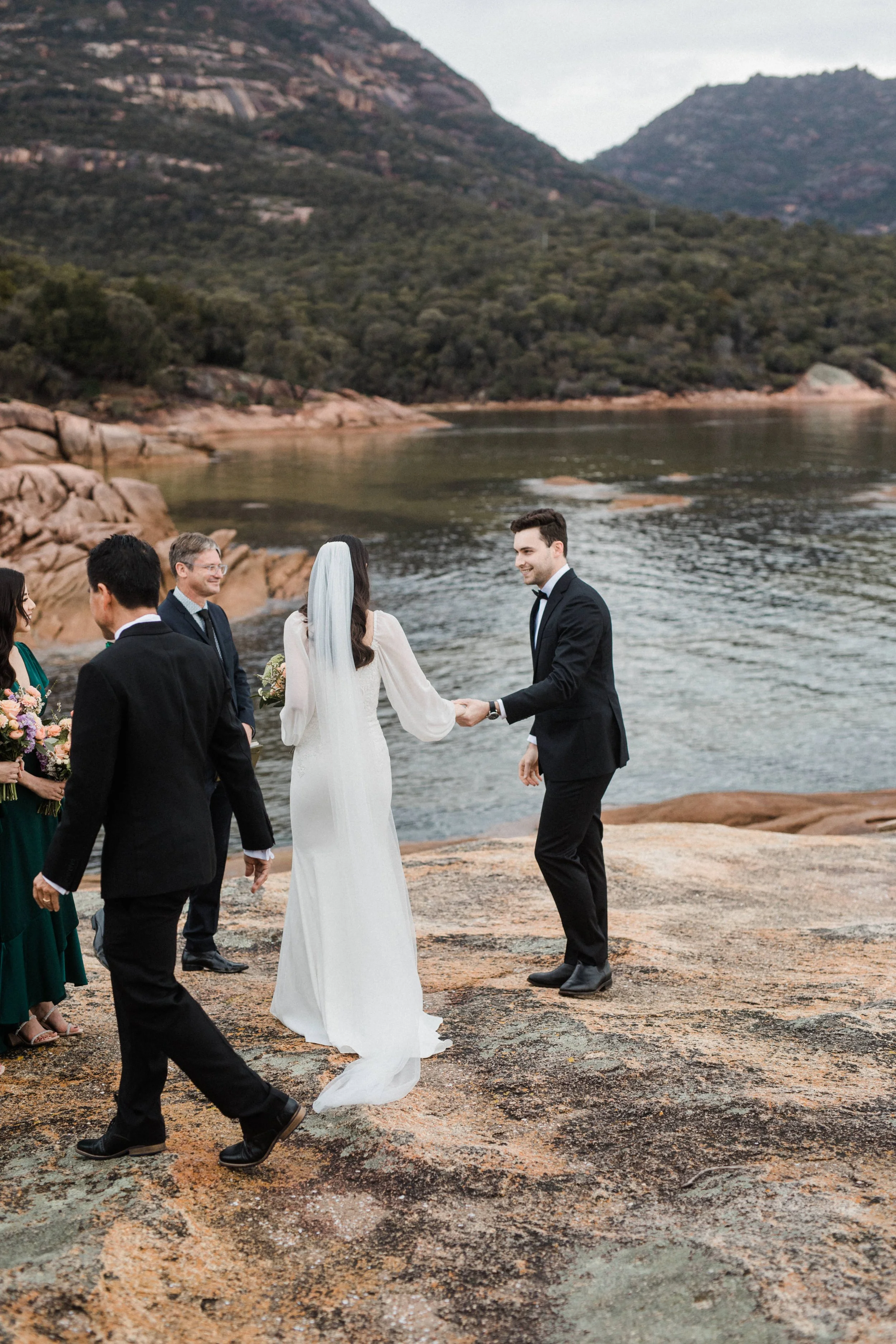 Bride and groom holding hands during an outdoor wedding ceremony by a river with mountains in the background.