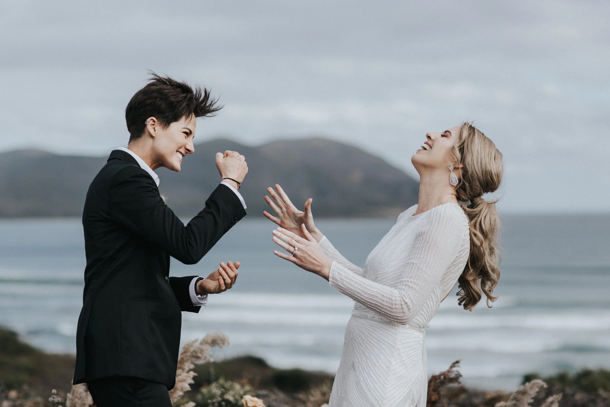 Two women laughing and celebrating at the beach, one in a white dress and the other in a black suit, with hills and the ocean in the background.