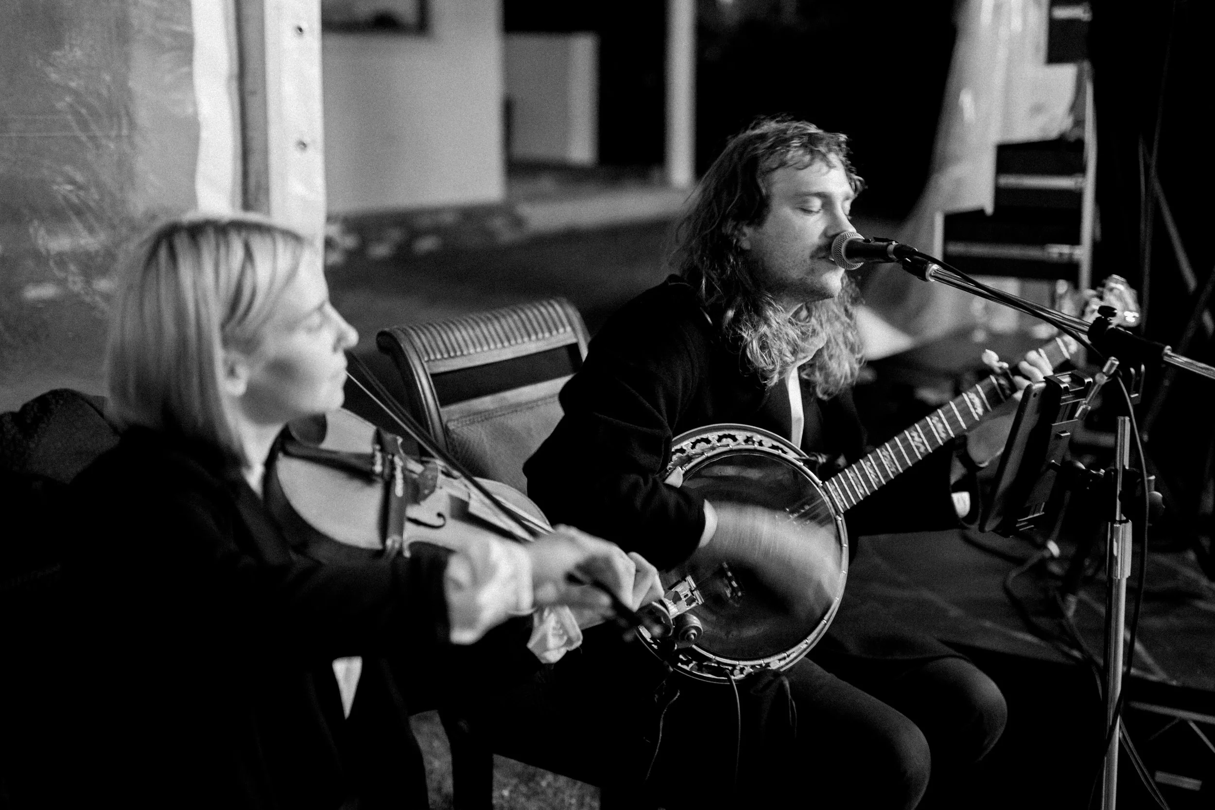 A black and white photo of a man and a woman performing music. The man is singing into a microphone and playing a banjo, while the woman is playing a violin.