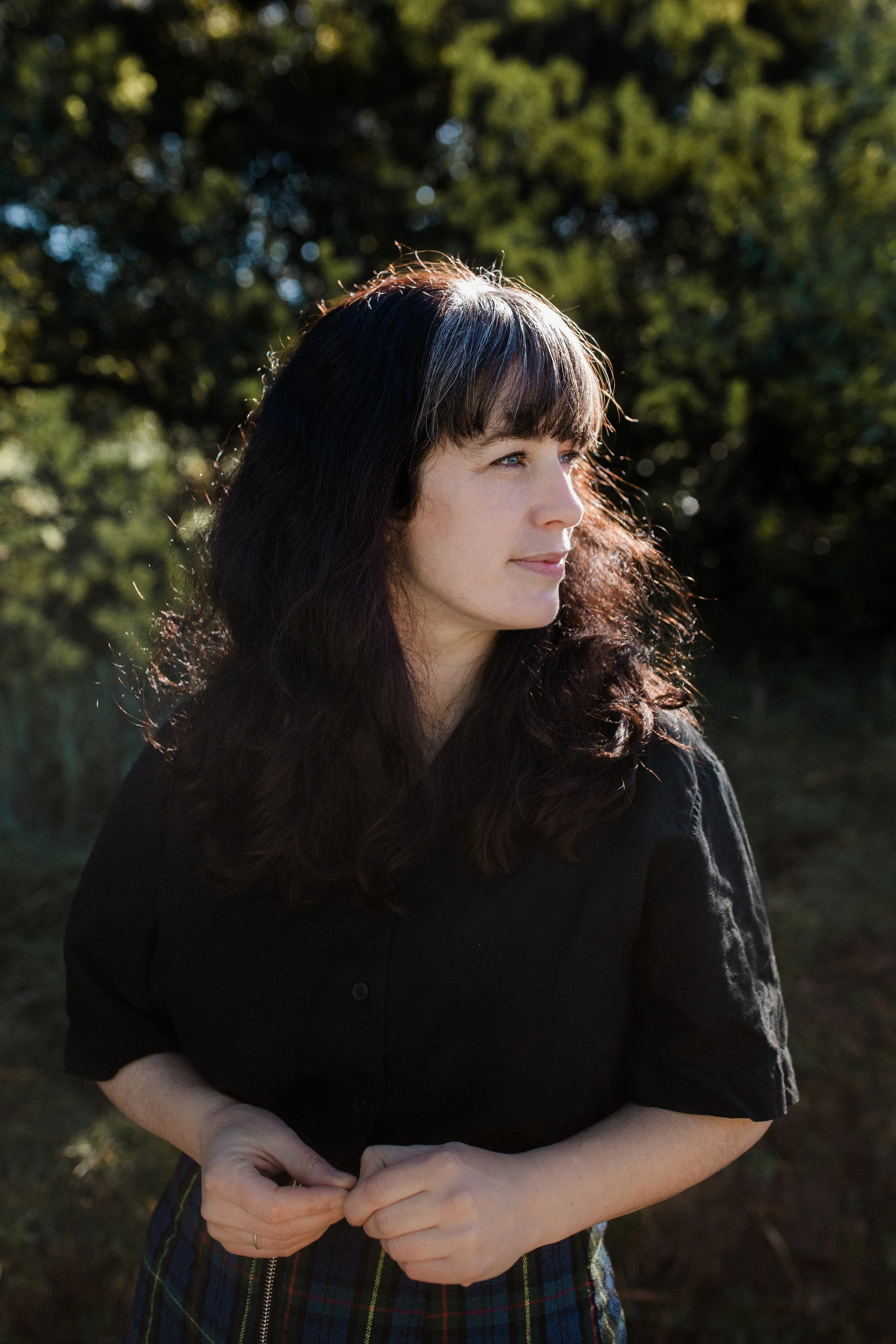 A woman with long dark hair and bangs standing outdoors in sunlight, wearing a black shirt and plaid skirt, with trees and greenery in the background.