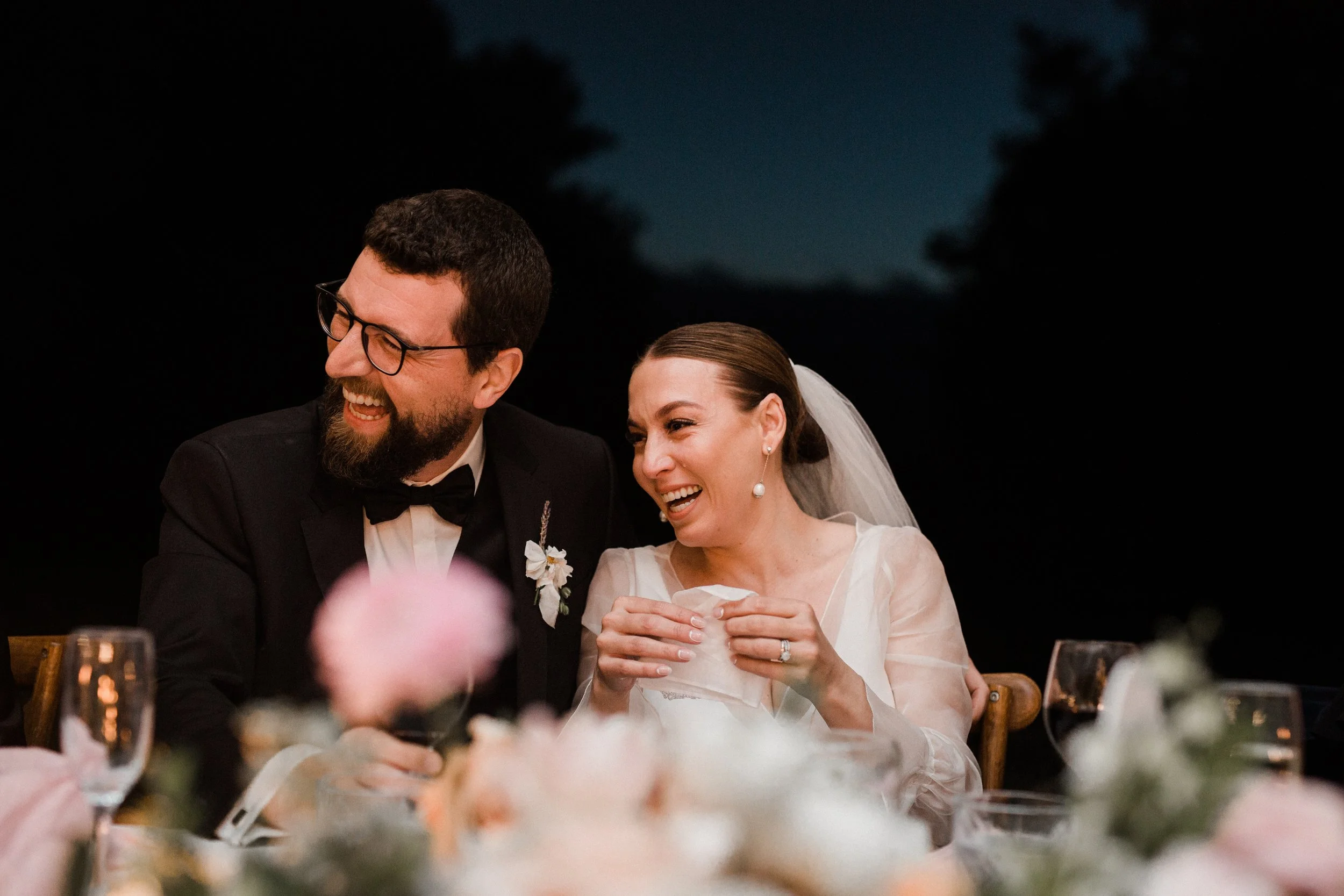 A bride and groom laughing and sharing a moment at their wedding reception indoors or outdoors at night, with floral decorations and glasses on the table.
