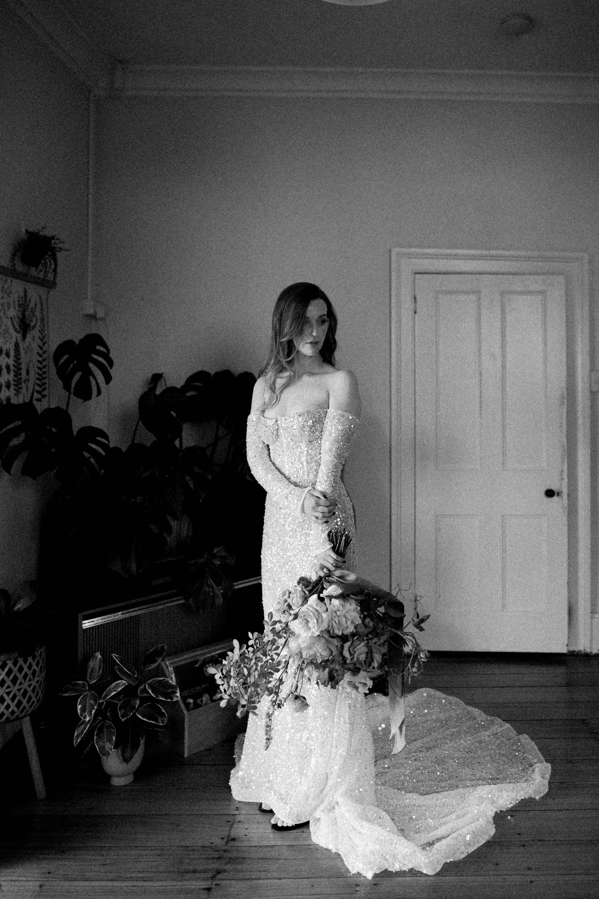 A woman in a sparkling gown holding a bouquet in a room filled with plants, standing in front of a closed door, in black and white photography.