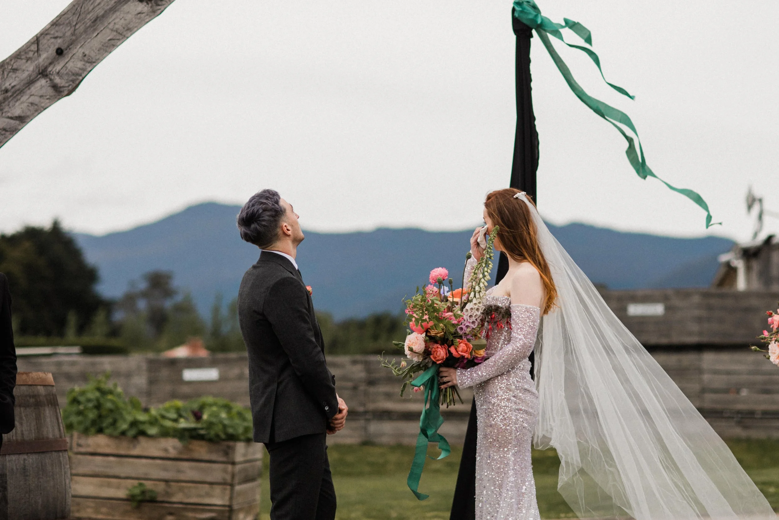Bride in a sparkly gown with a long veil holding a bouquet of pink and orange flowers, standing in front of groom in a suit, during an outdoor wedding ceremony with mountain scenery in the background.