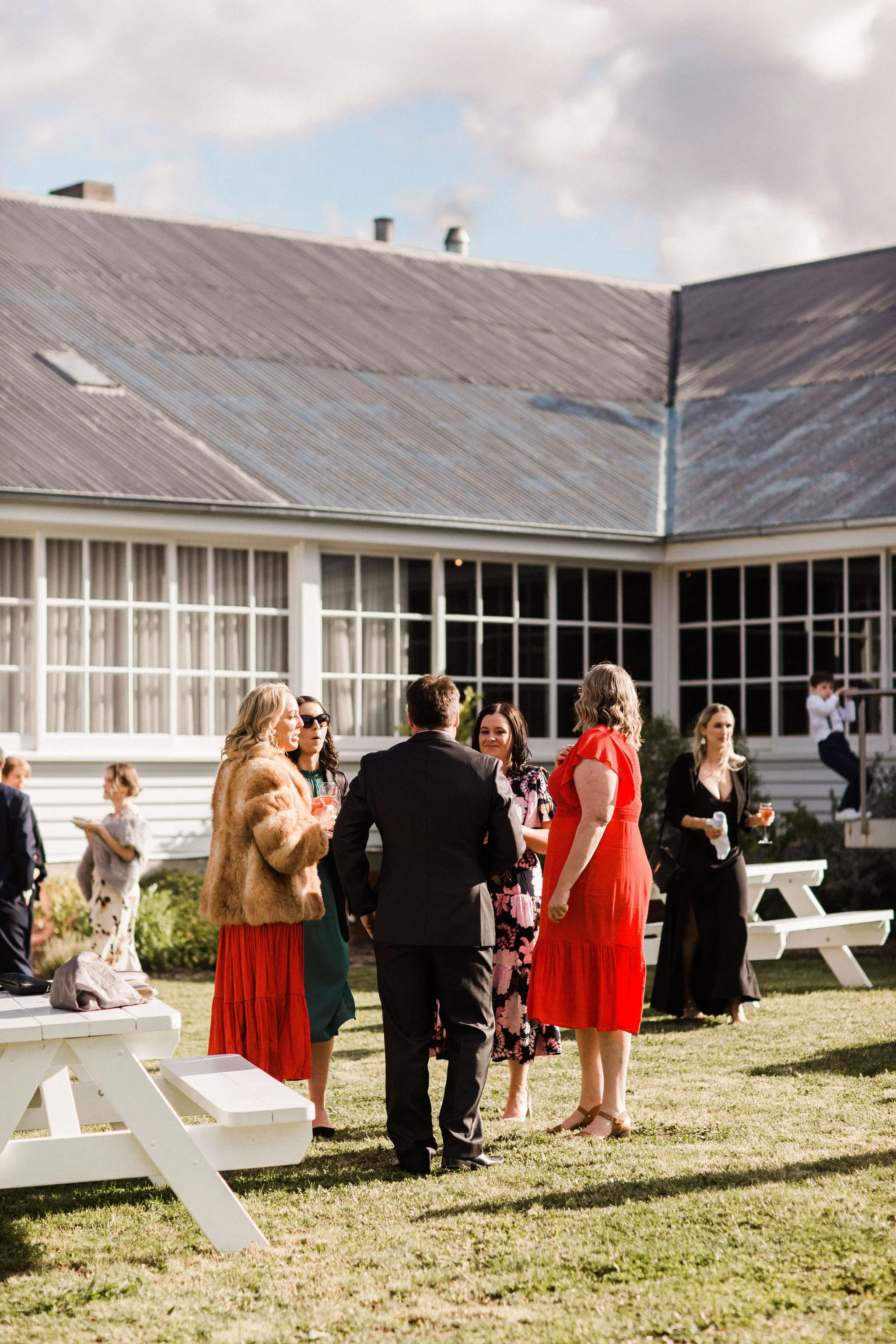 A group of people socializing outdoors at a gathering, with some dressed in formal and semi-formal clothing, near a white building with large windows, on a sunny day.
