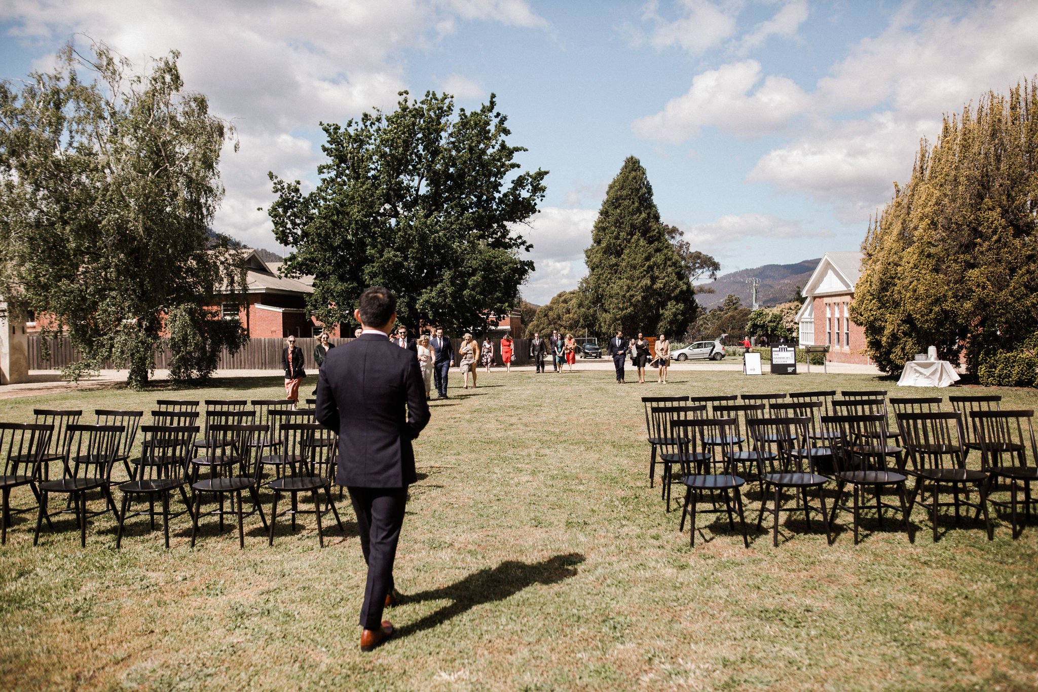 Man in a black suit walking towards an outdoor wedding ceremony setup with chairs arranged on either side of the aisle, guests in formal attire gathering in the background, trees, a building, and mountains under a partly cloudy sky.