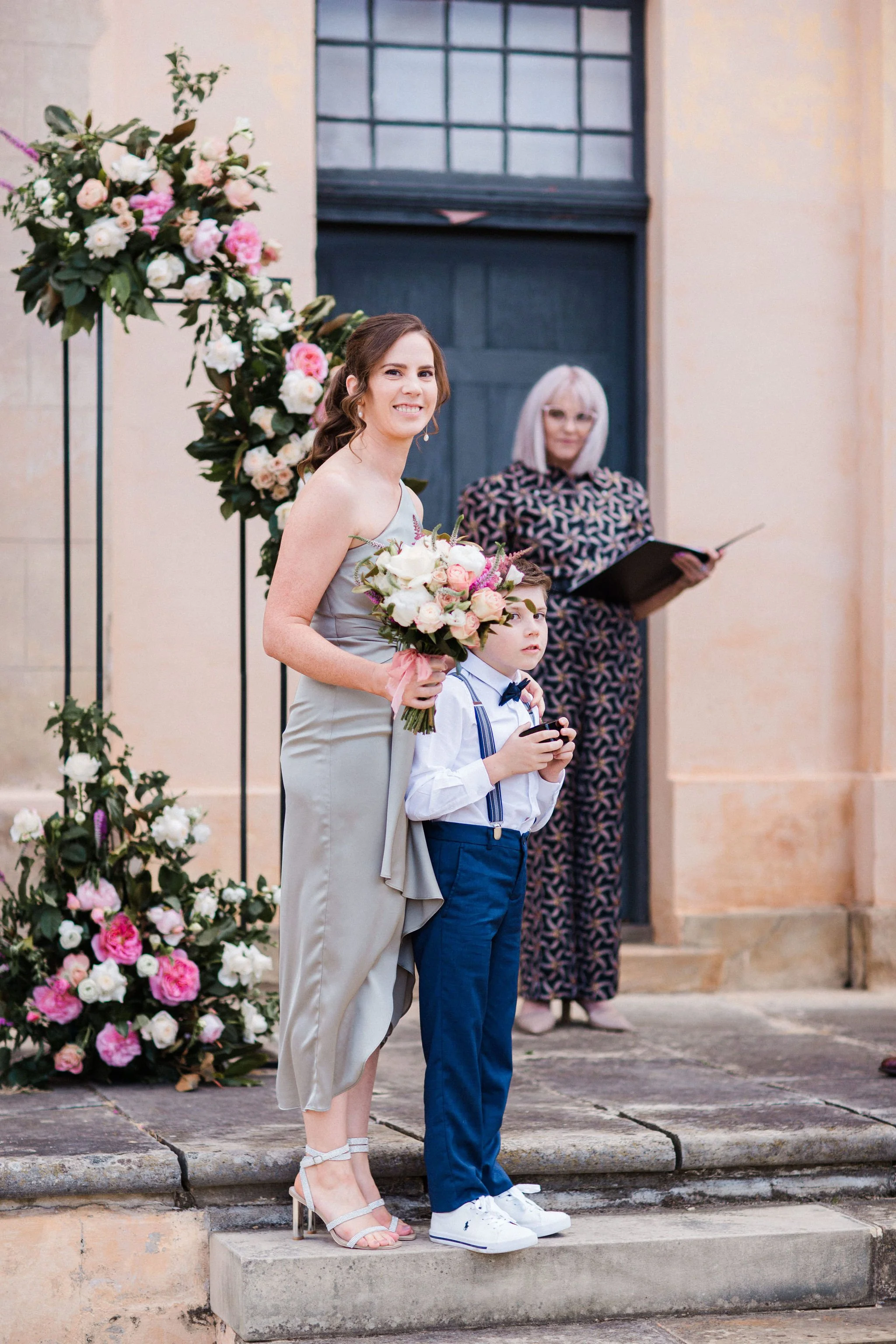 A woman in a light gray dress holding a bouquet of flowers standing next to a young boy in a white shirt, blue pants, and suspenders with a bow tie, at an outdoor wedding ceremony. A woman with blond hair holding a black folder is in the background i