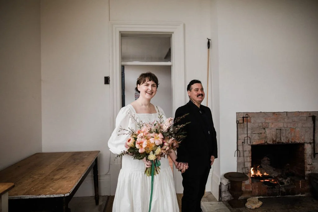 A bride and a man holding hands, standing indoors near a fireplace, smiling. The bride is wearing a white dress and holding a large bouquet of pink and peach flowers. The man is dressed in black.