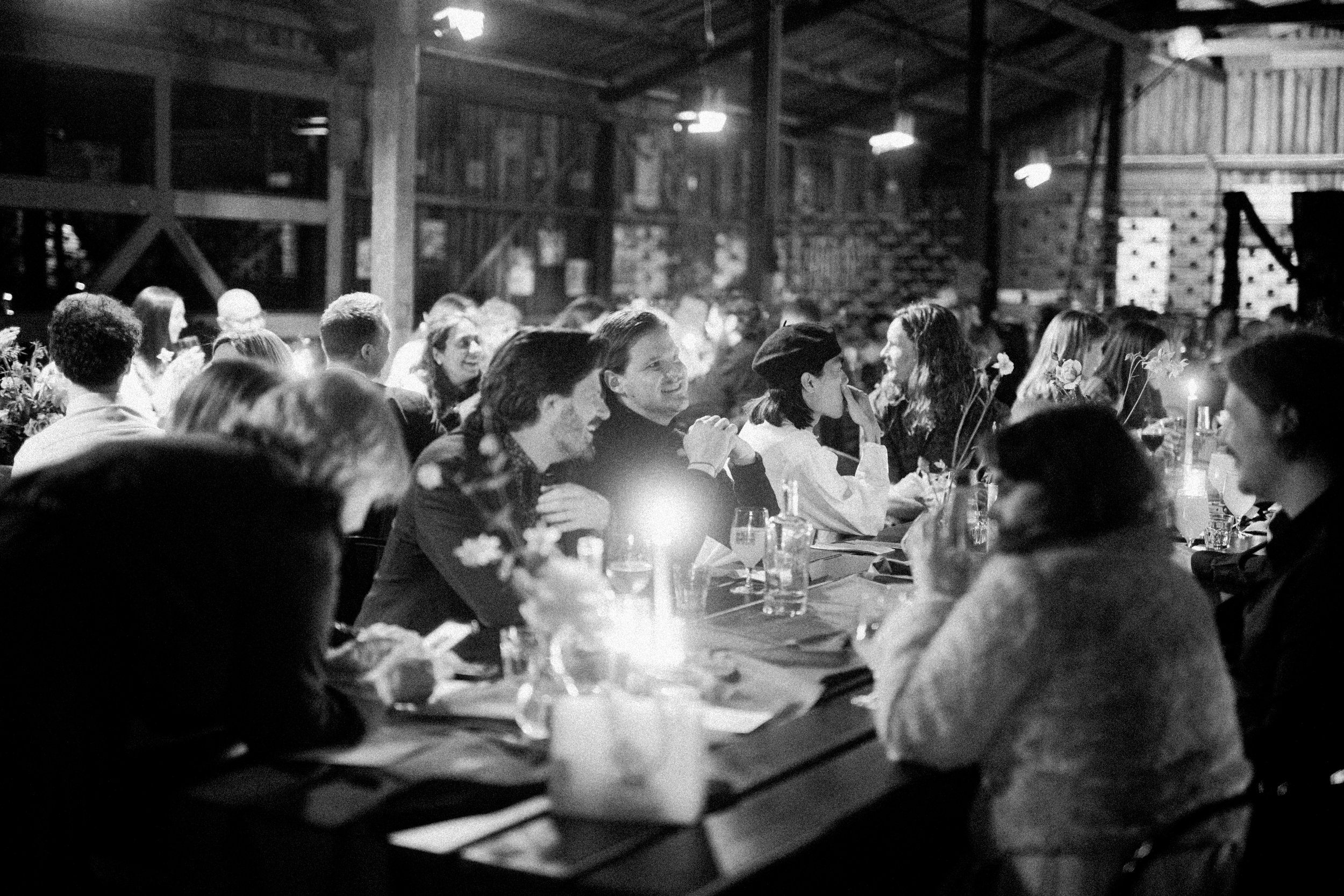 People seated at a long table, laughing and conversing, in a rustic barn setting with wooden walls and soft lighting.