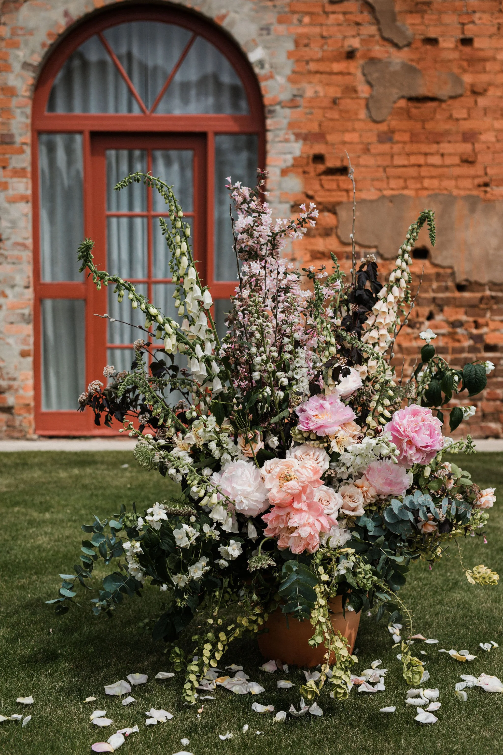 A large floral arrangement in a terracotta pot featuring pink and white flowers, sitting on a lawn with scattered petals. In the background, there is a red brick building with a large arched window with curtains.