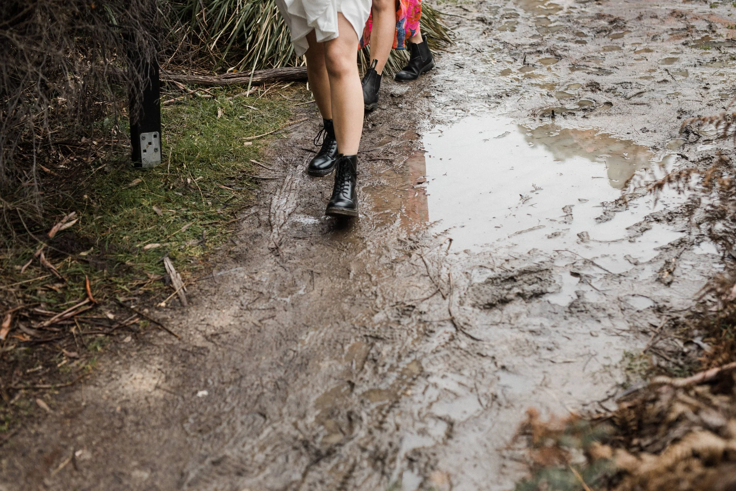 Close-up of two people walking on a muddy trail in wet weather, wearing black boots and colorful clothing.