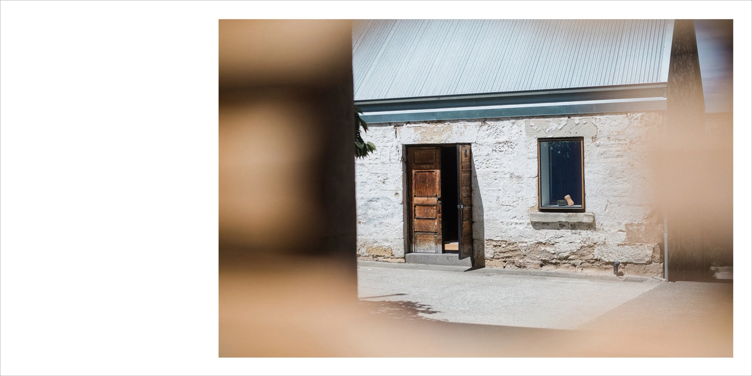 The image shows the exterior of a rustic stone house with a wooden door that is slightly open, and a window with a small object on the windowsill. The photo is taken through an opening or some blurry foreground elements, giving a framed view of the h