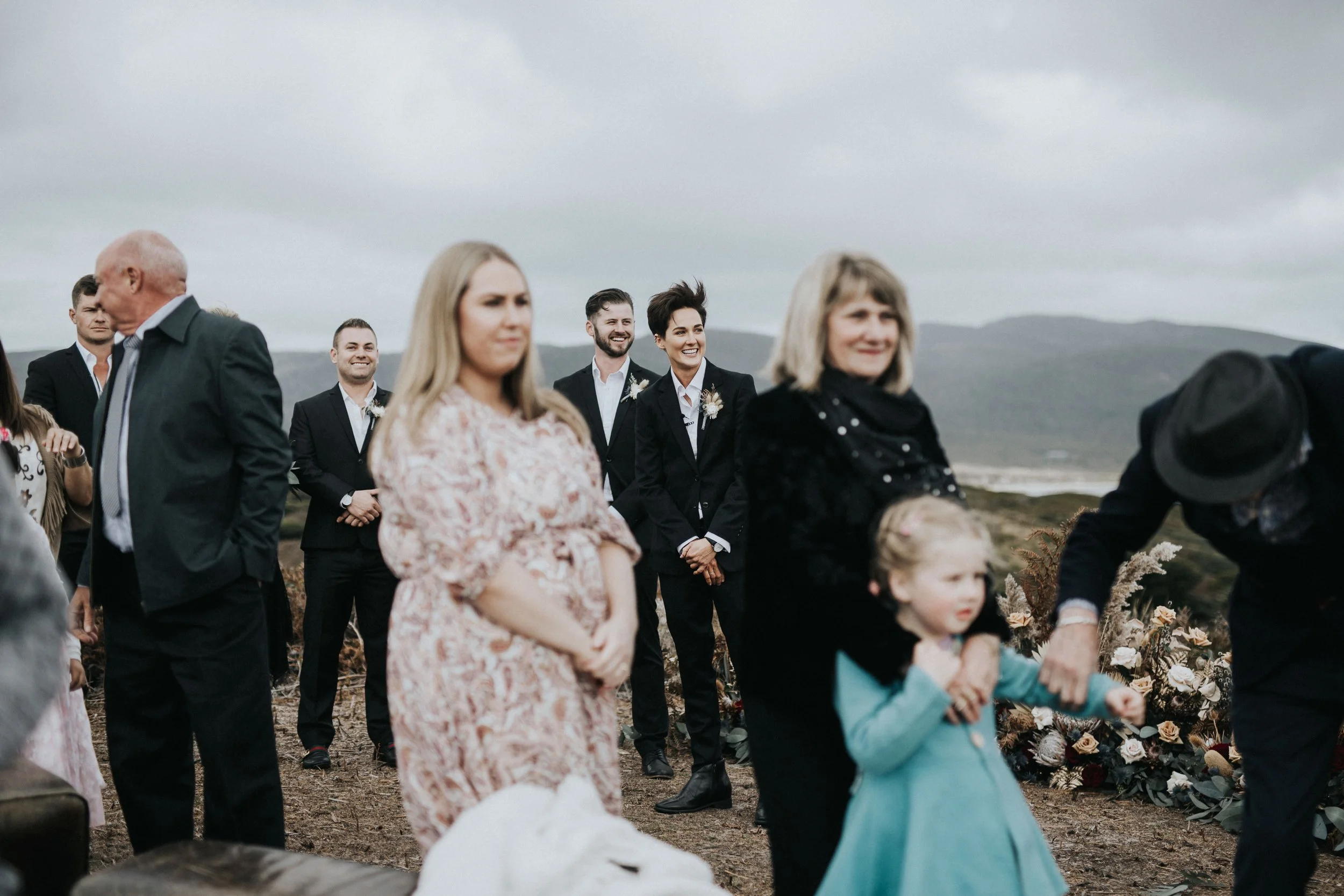 People attending an outdoor wedding ceremony on a cloudy day, with a backdrop of hills and the ocean, including women, men, and a child.