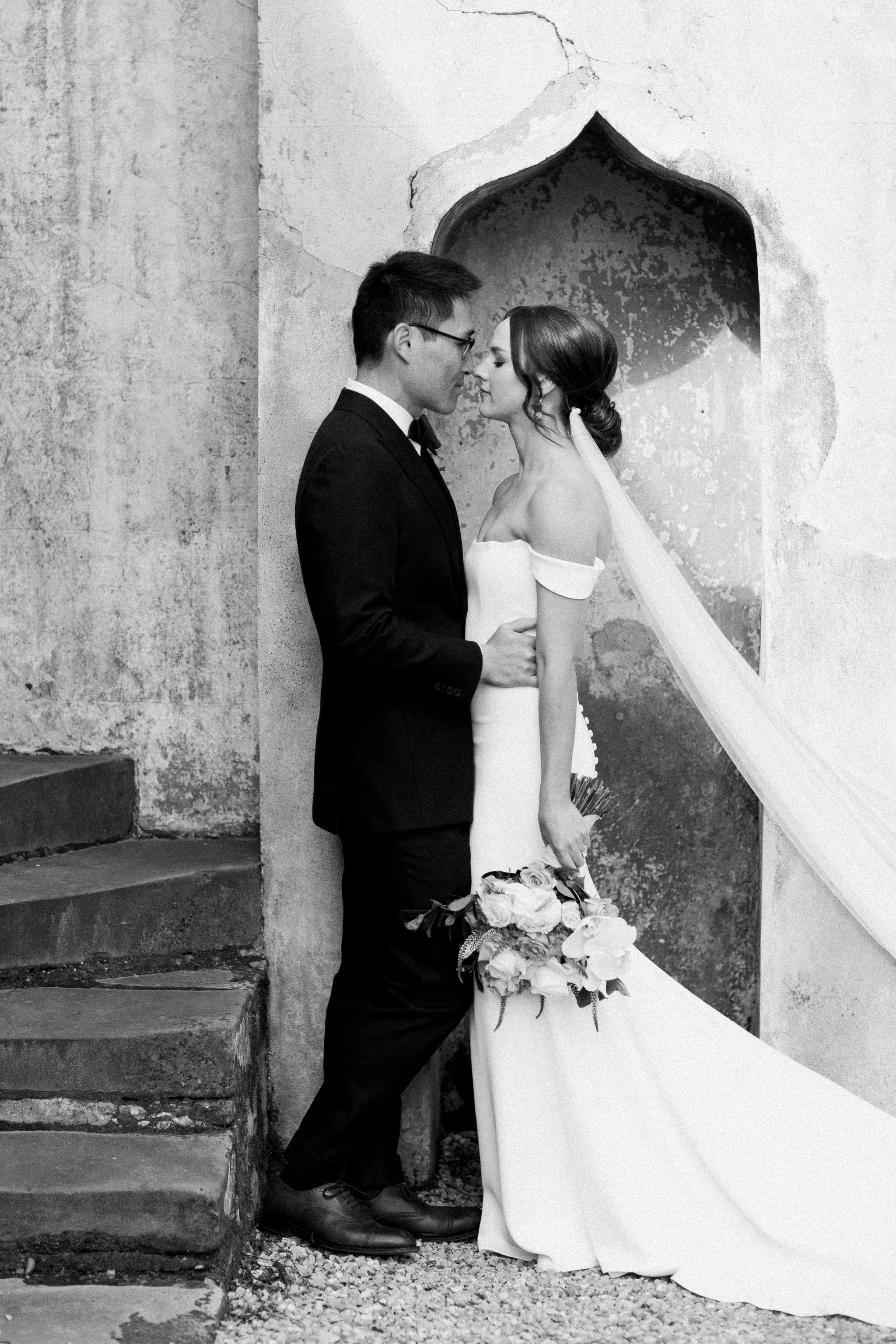 A black and white photo of a bride and groom near a textured wall with an arched opening. The groom wears a suit and glasses, the bride wears an off-shoulder wedding dress and holds a bouquet, standing closely with foreheads touching.