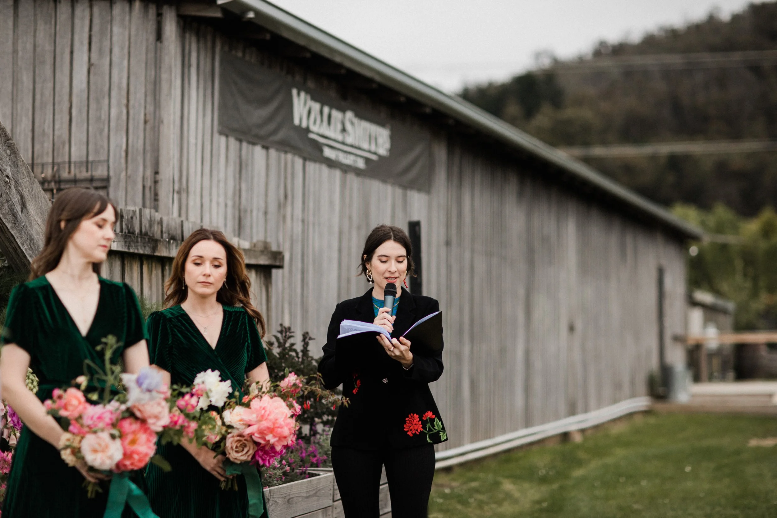A woman with short dark hair and earrings is speaking into a microphone while reading from a notebook. She is dressed in a black blazer with floral embroidery. To her left, two women with long brown hair, wearing dark green dresses, hold bouquets of 