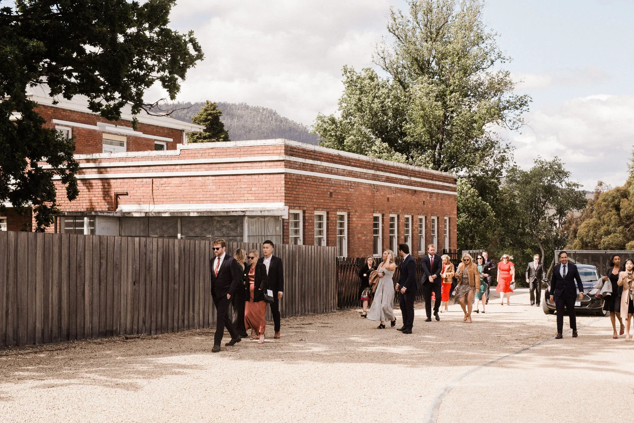 Group of people dressed in formal attire walking outdoors on a sunny day near a brick building with trees in the background.