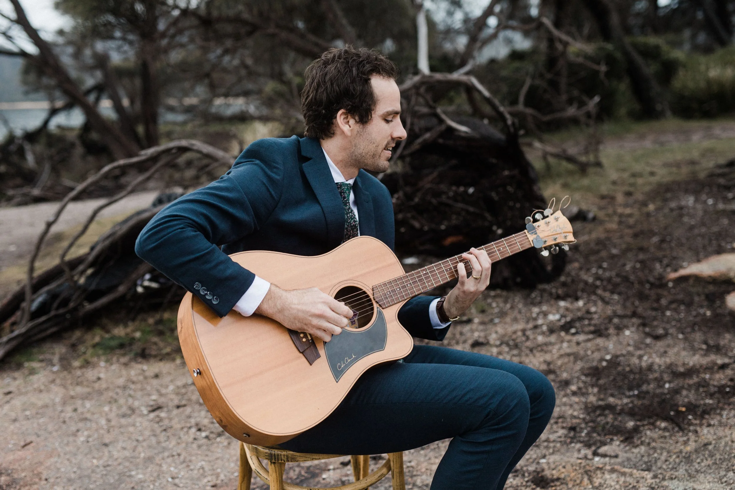 A man in a dark blue suit playing an acoustic guitar outdoors, sitting on a wooden stool near a fallen tree.
