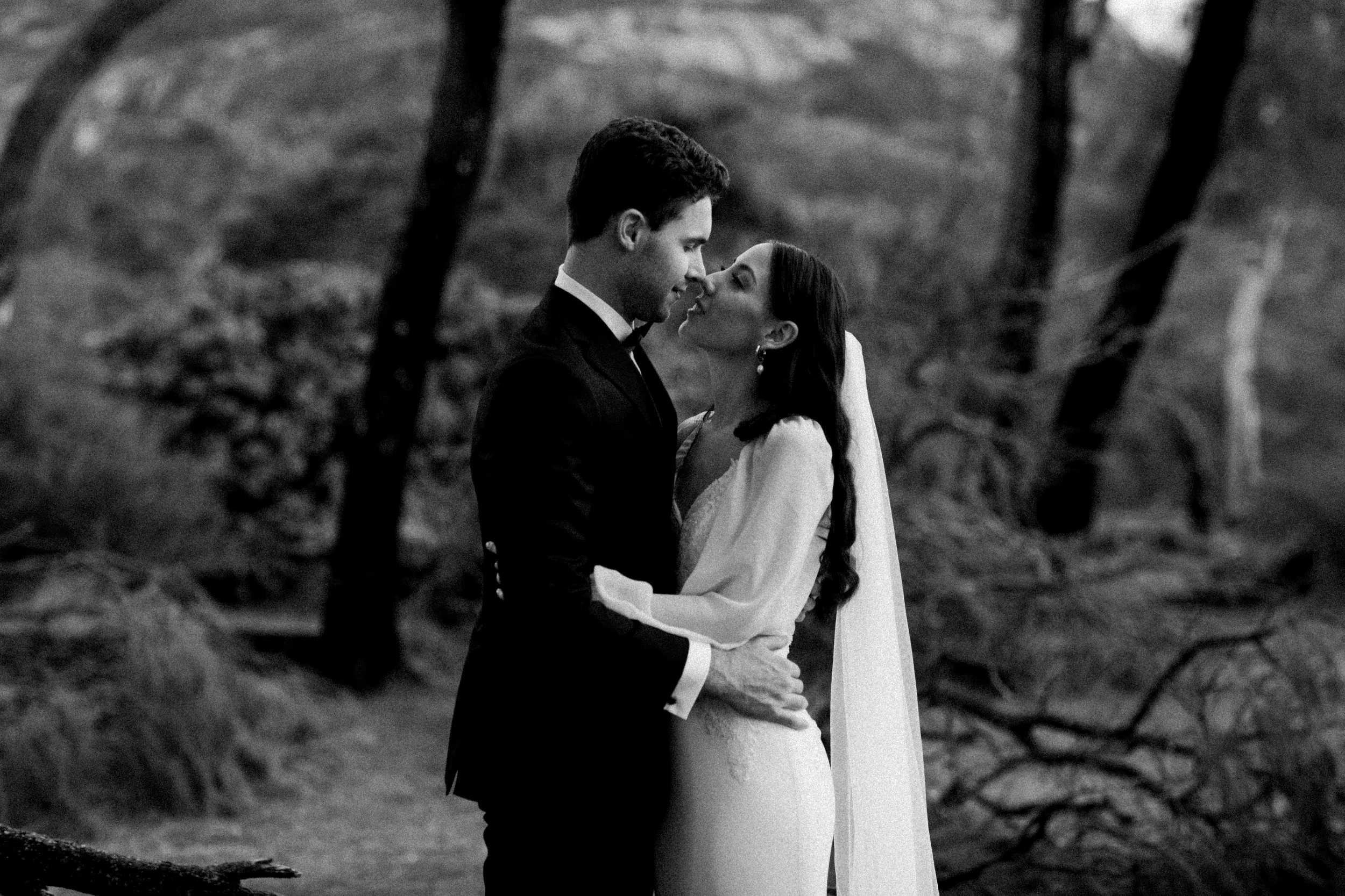 A black and white photo of a bride and groom embracing outdoors, close to each other, about to kiss, with trees in the background.