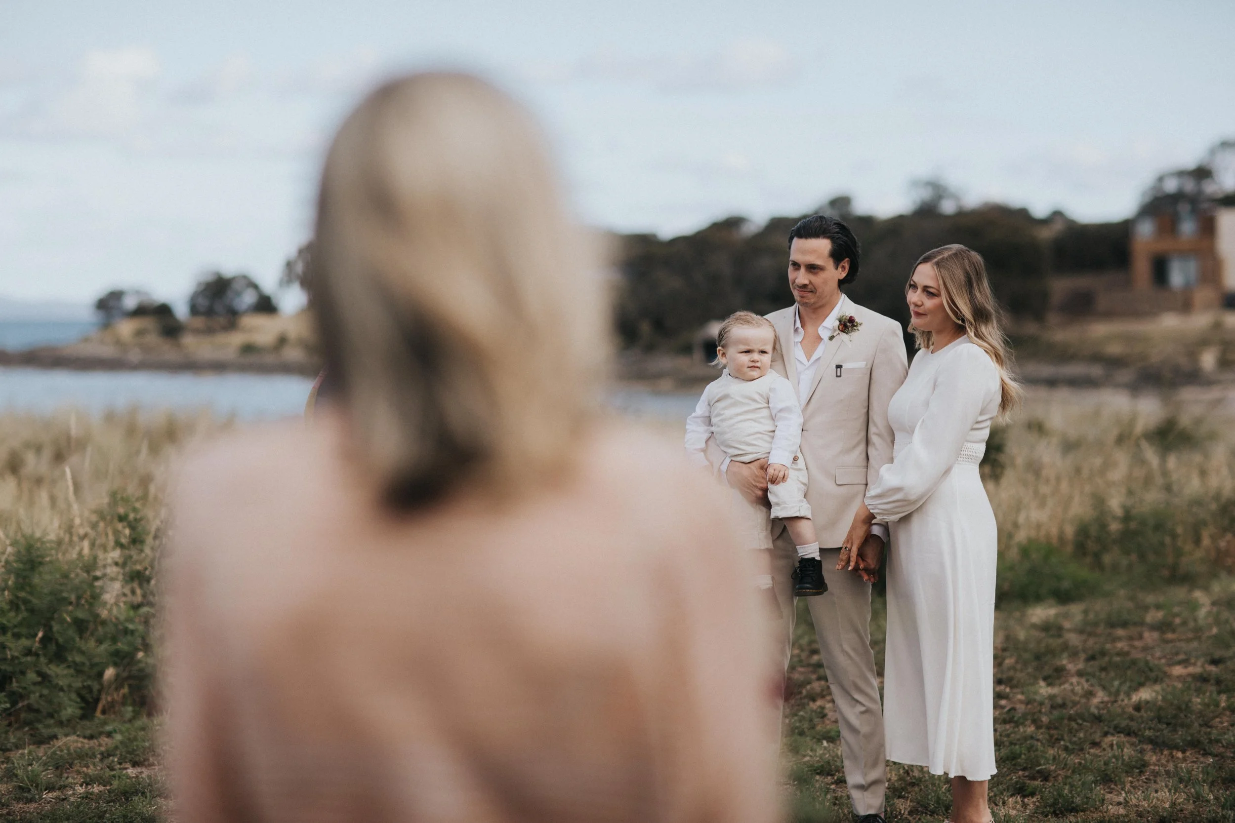 A couple with a child at a beachside wedding ceremony, with the woman in white and the man in beige suit holding a young boy.