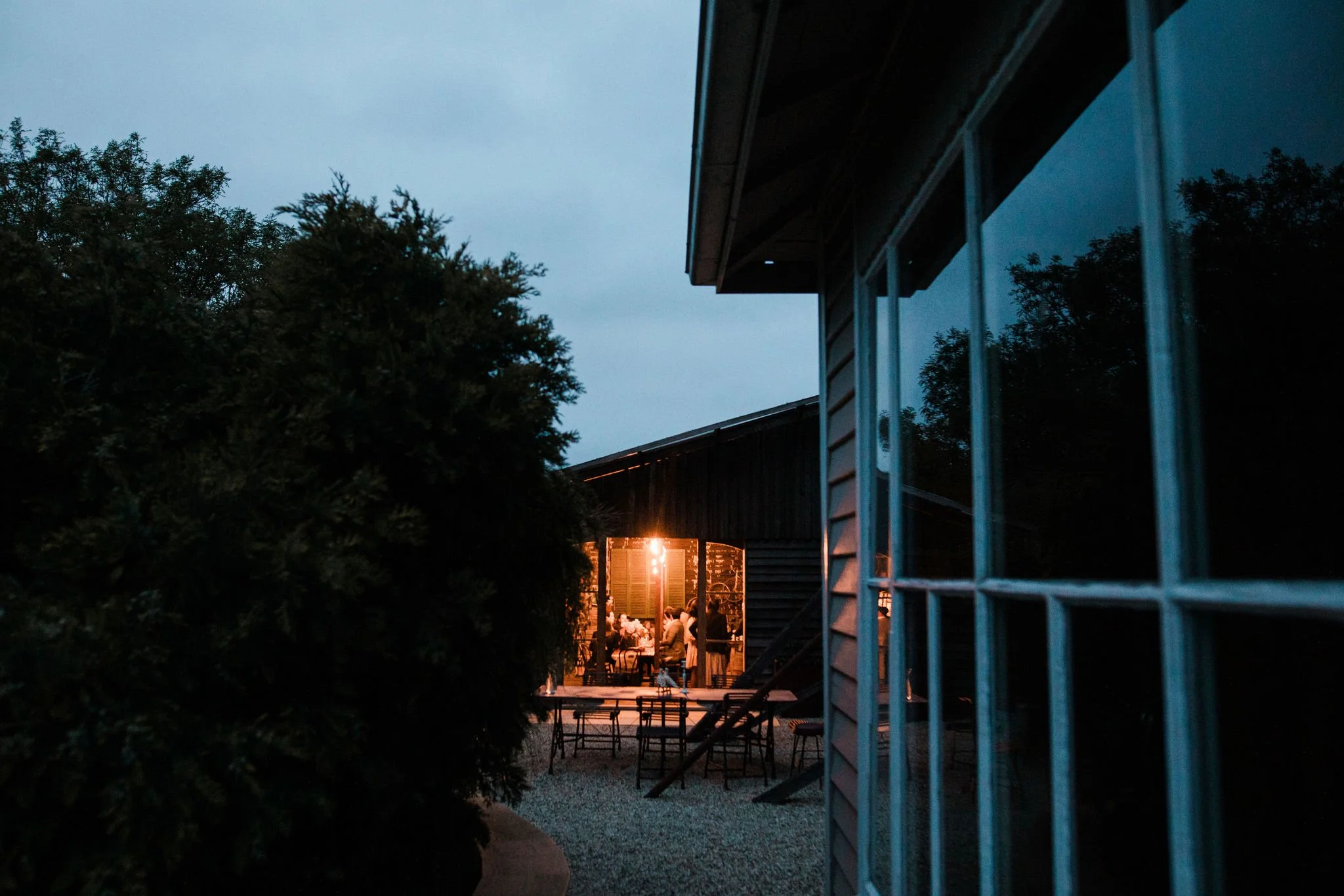 An evening outdoor gathering with people under string lights, viewed from outside a house through a window and bushes.
