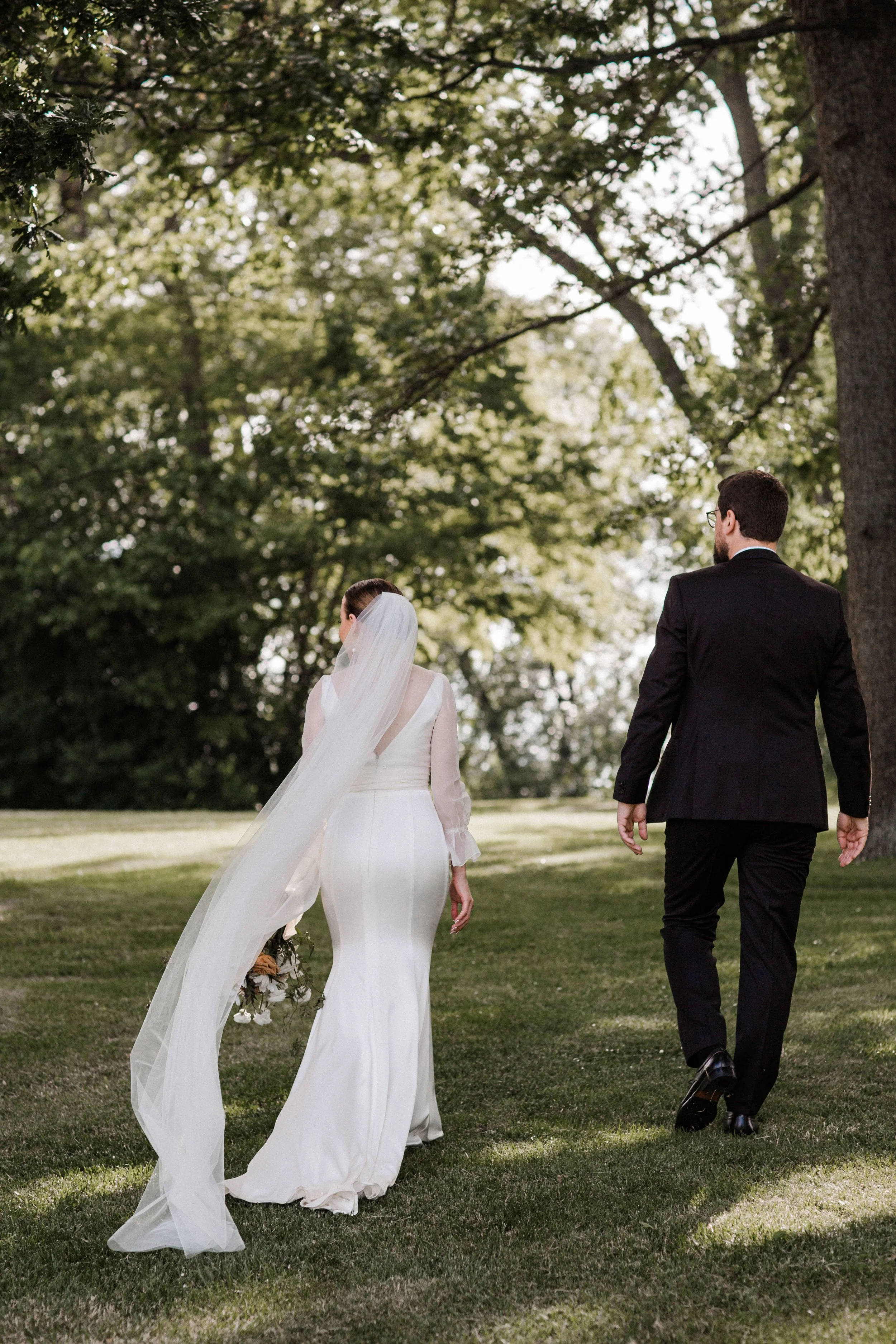 A bride and groom walking together outdoors on a grassy area under tall trees, seen from behind during a wedding ceremony.