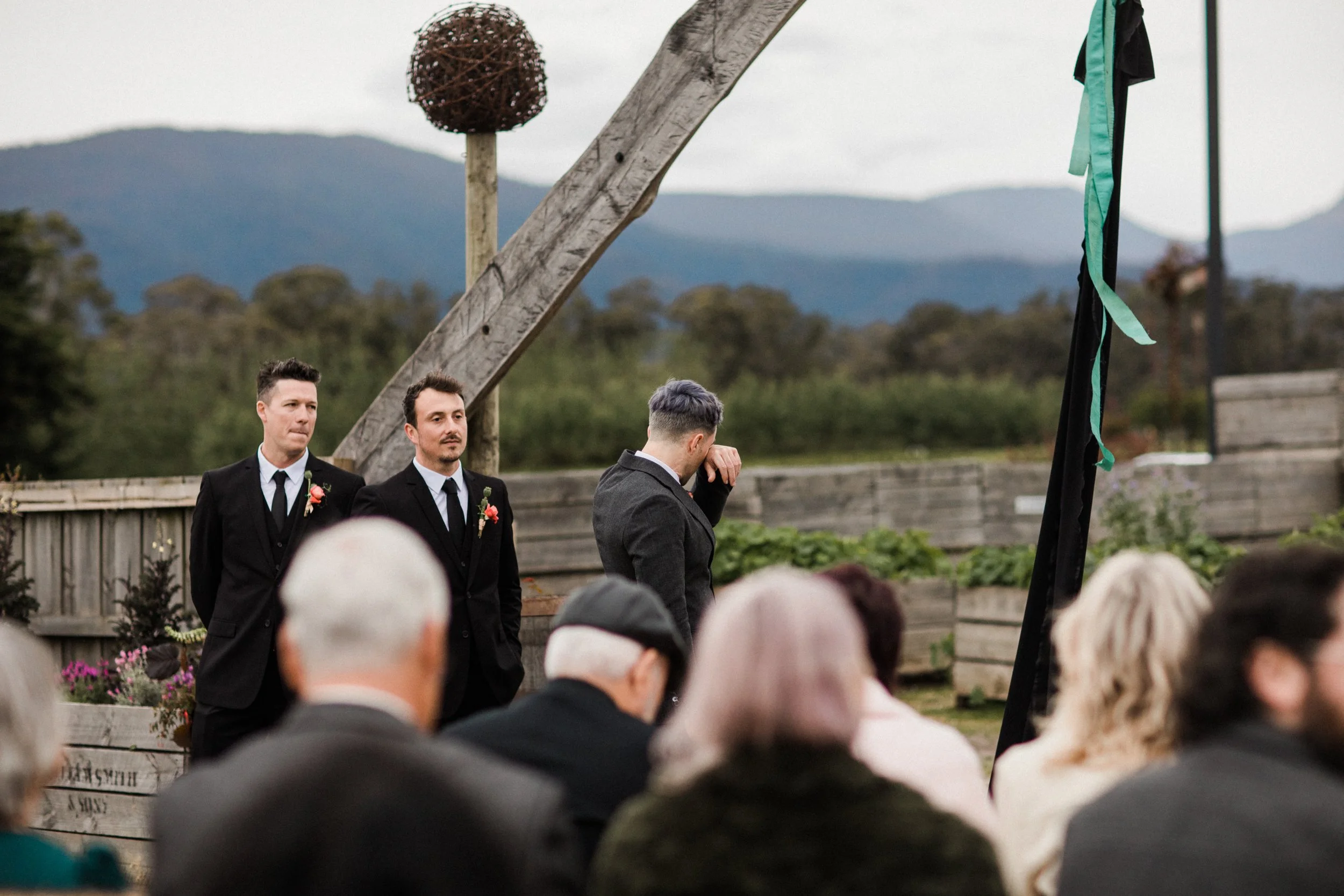 An outdoor wedding ceremony with three men dressed in suits standing in front of an audience, with one man wiping a tear, against a scenic mountain backdrop.