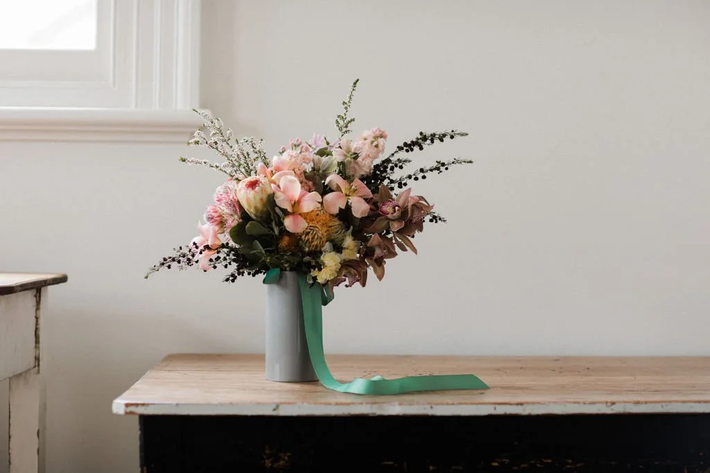 A bouquet of pink, white, and purple flowers in a gray vase with a teal ribbon, placed on a wooden table against a plain wall.
