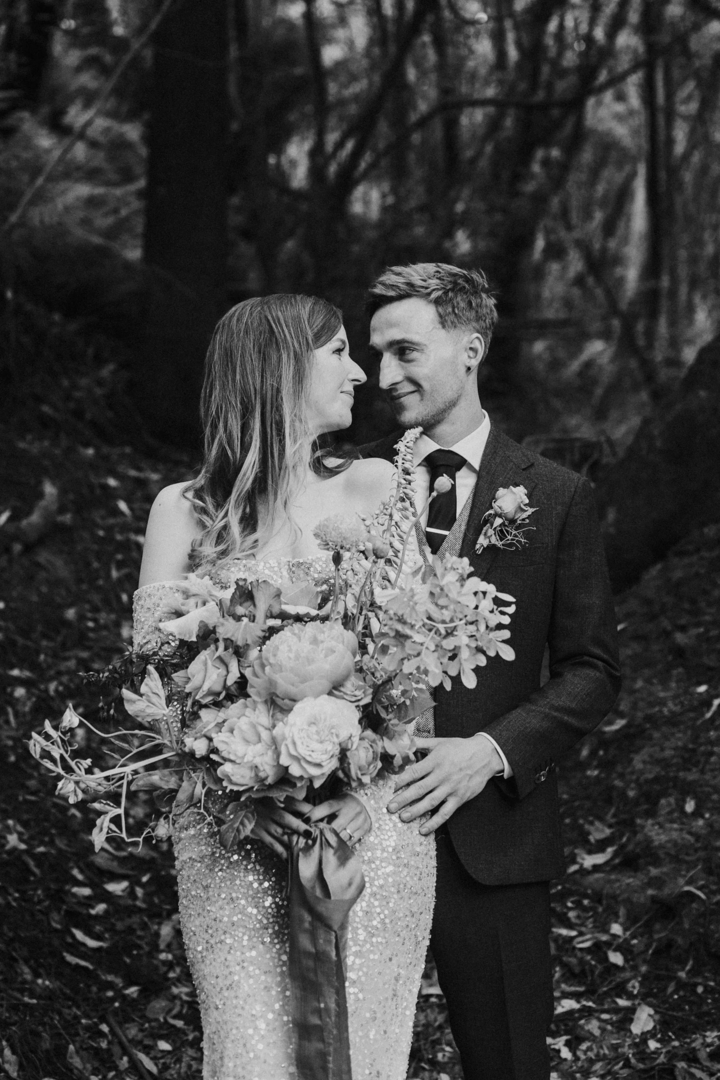 A black and white photo of a bride and groom in a forest, gazing into each other's eyes. The bride is holding a large bouquet of flowers and wearing a sparkly dress, while the groom is dressed in a dark suit with a boutonnière.