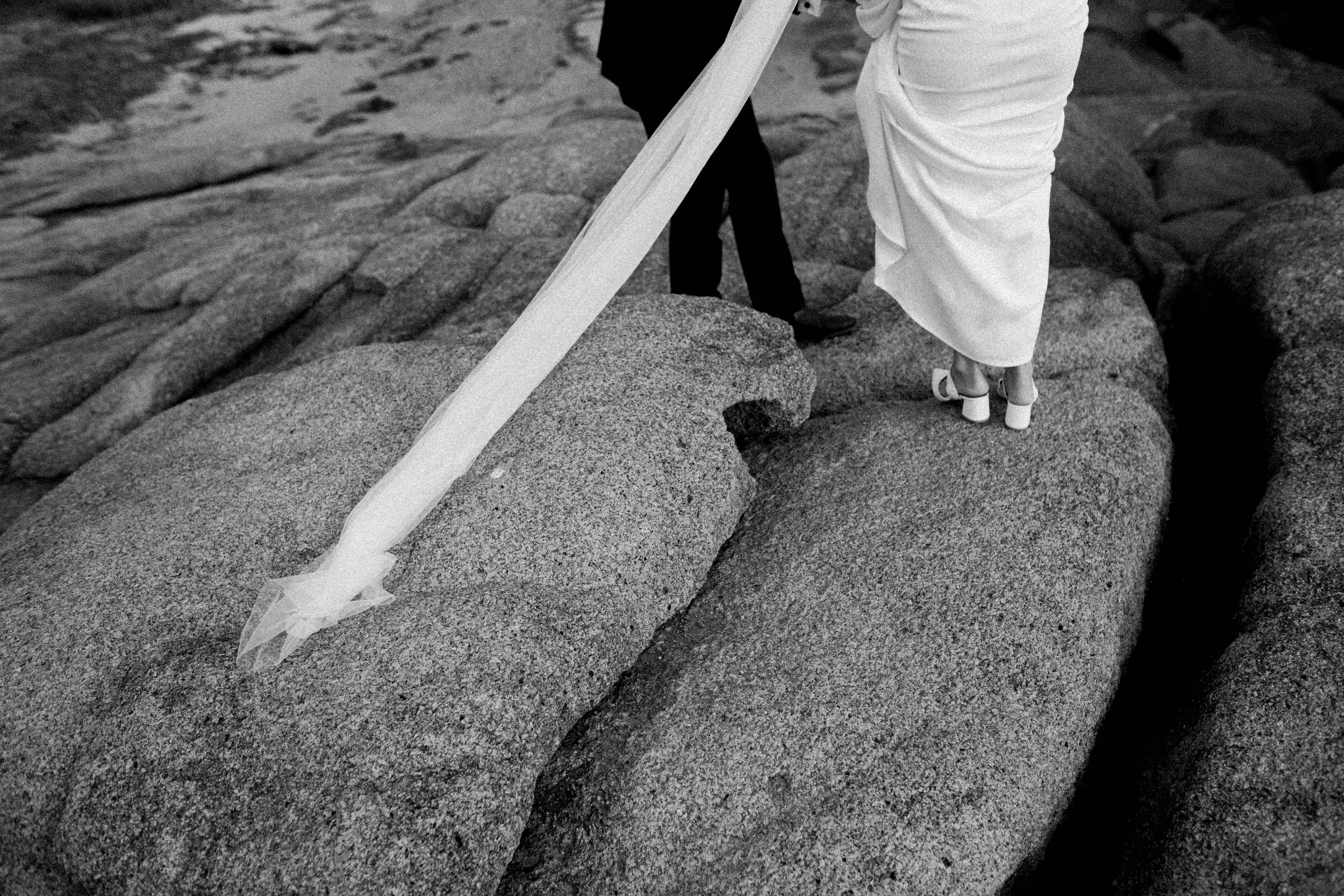 A woman in white heels and a white dress, possibly a wedding gown, walking on large rocks near water, with a man in dark clothing standing nearby.