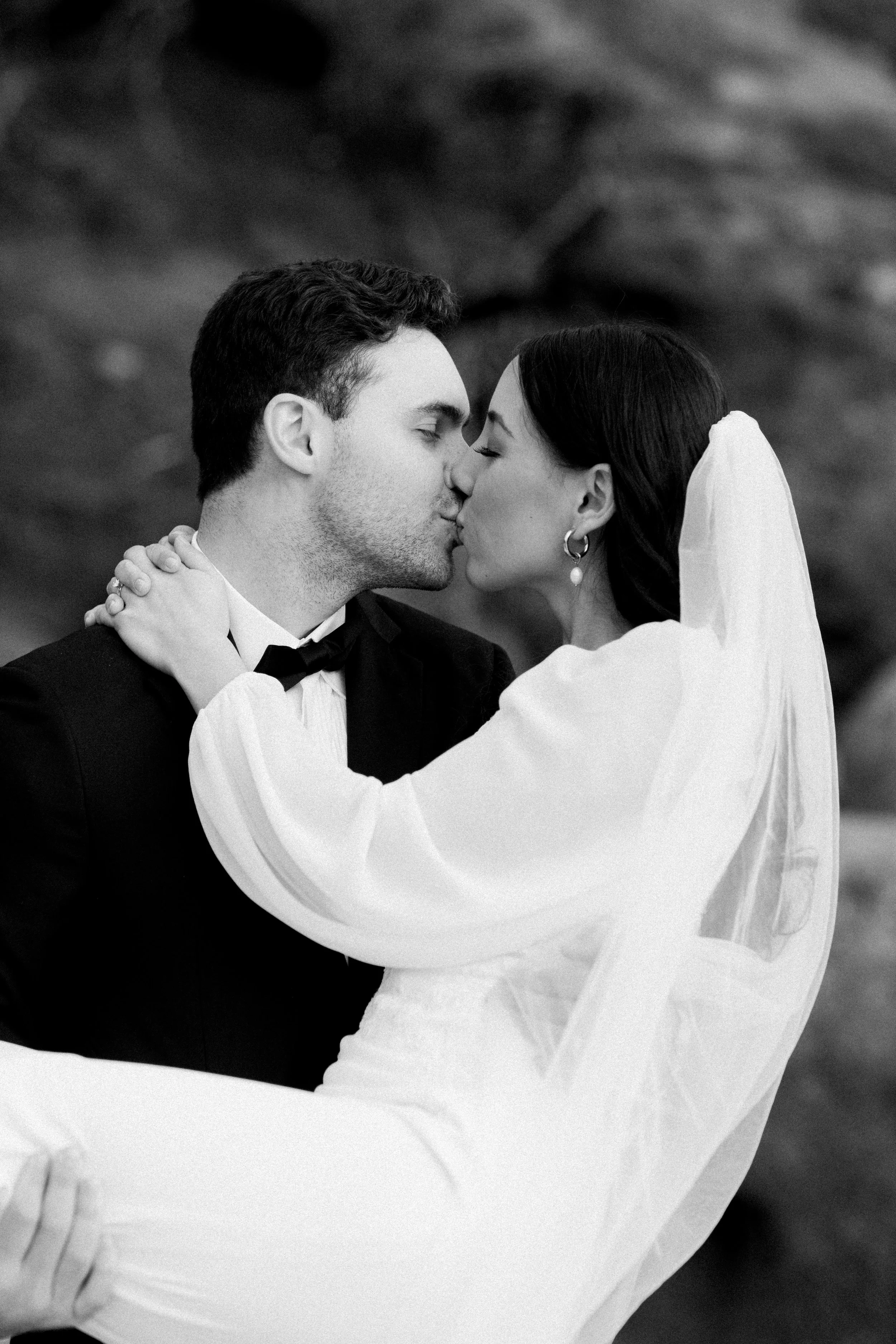 A black and white photo of a bride and groom sharing a kiss, with the groom in a tuxedo and the bride in a wedding dress and veil.