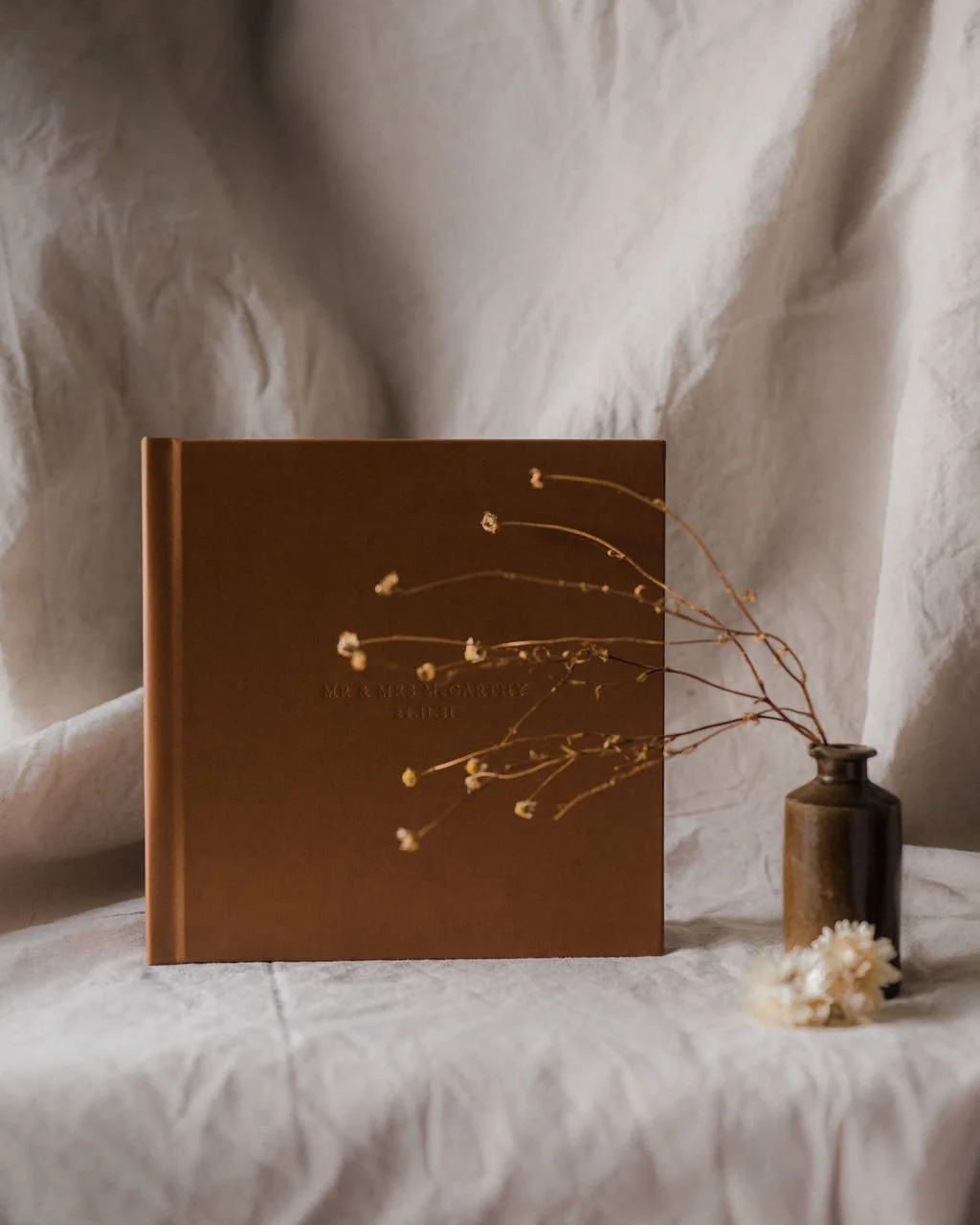 A brown hardcover book with floral illustrations on the cover, placed next to a small brown vase with dried flowers, all set against a crumpled fabric backdrop.