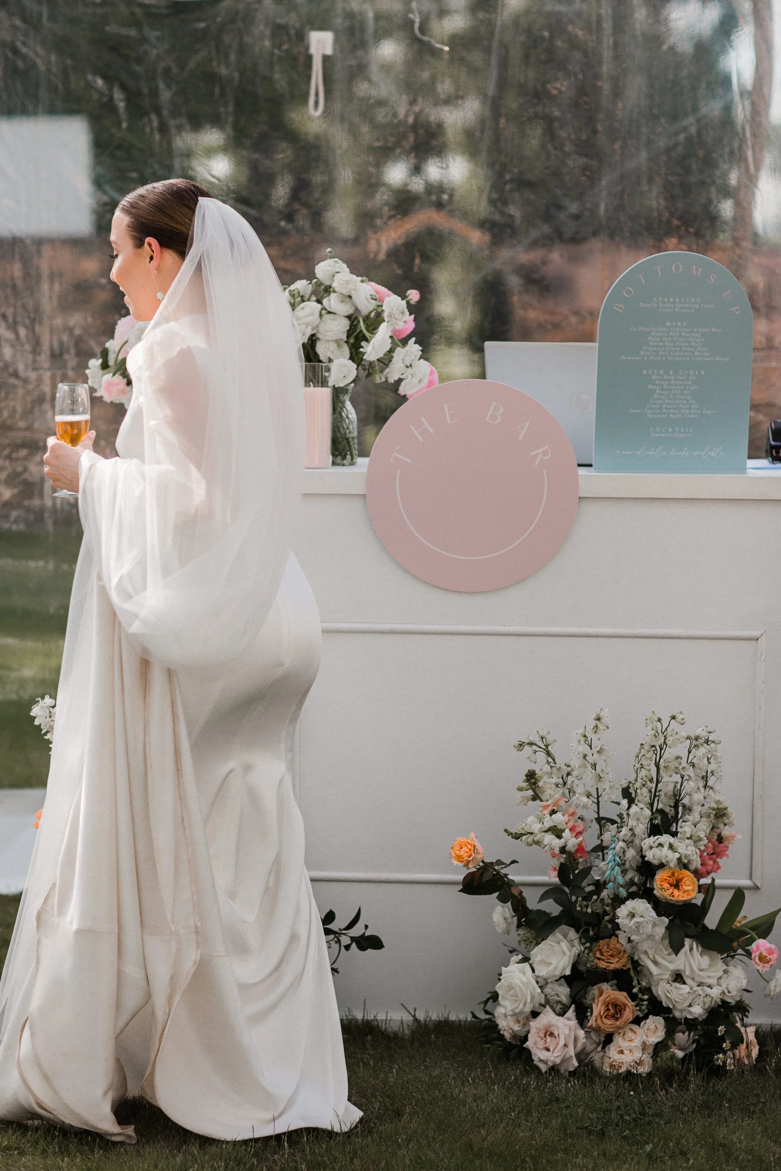 A bride in a white wedding dress and veil standing next to a white bar station holding a glass of rosé wine, with floral arrangements and a menu sign in the background.