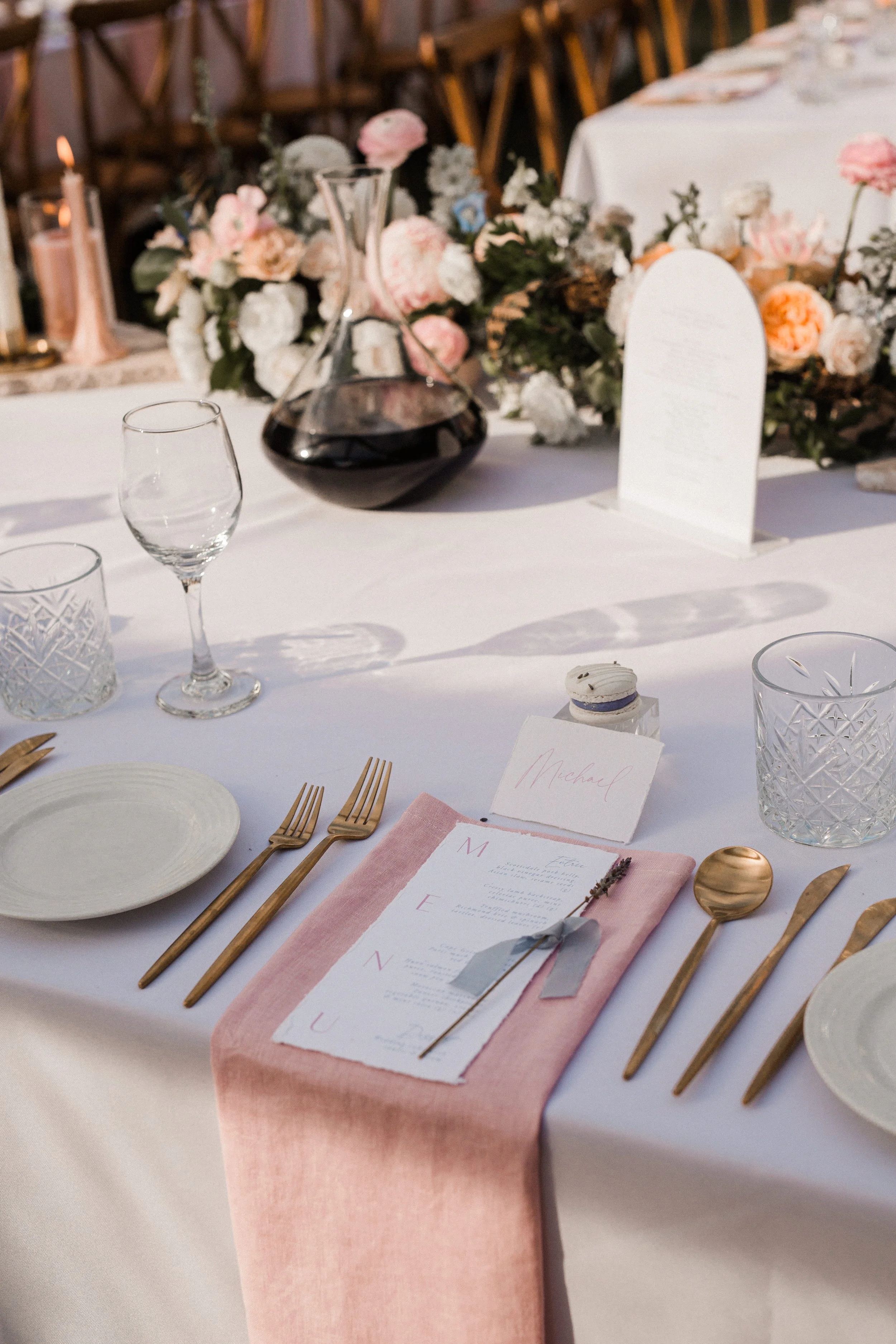 Wedding reception table set with white tablecloth, pink and gold accents, floral centerpiece, wine decanter, glassware, menu card, and place card.