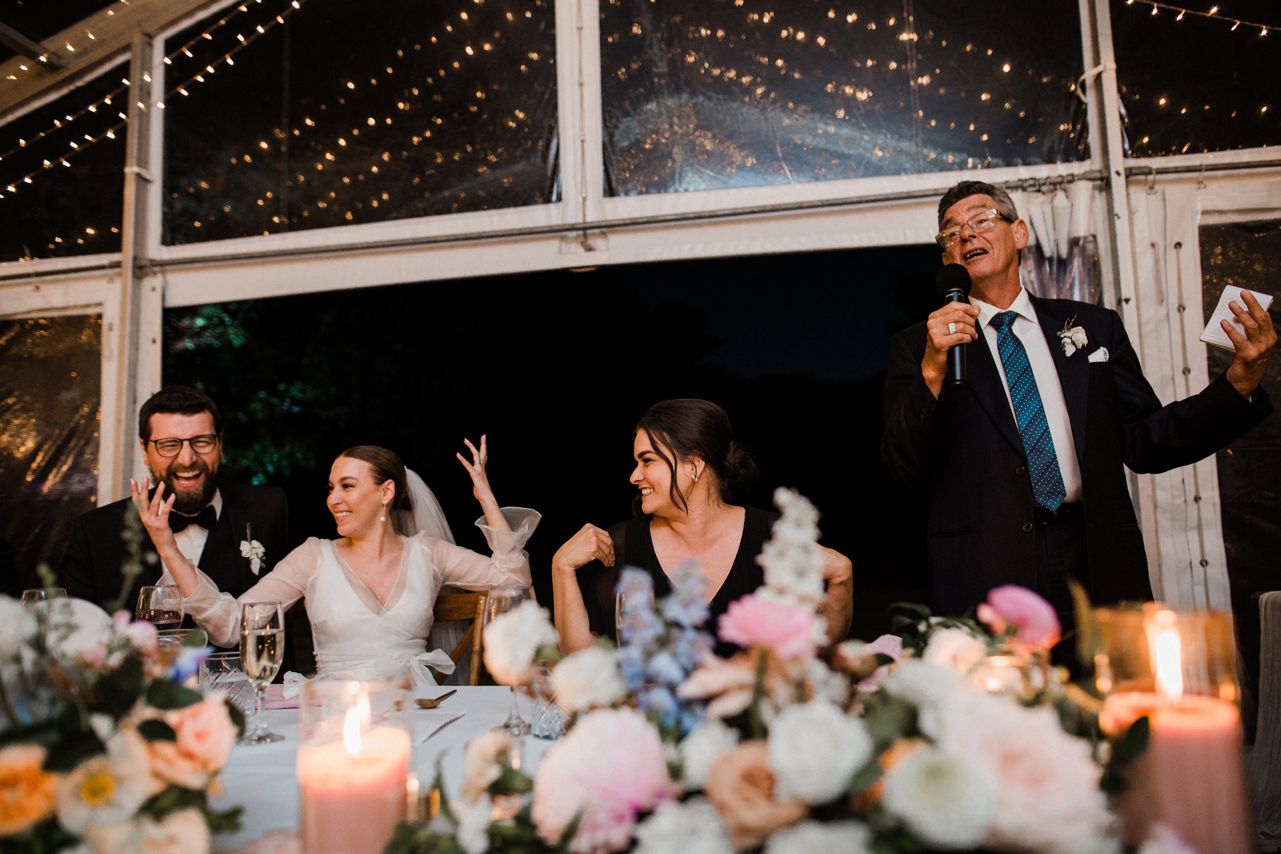 At a wedding reception, four people sit at a decorated table with flowers and candles. A man in a tuxedo is giving a speech, holding a microphone with a smile. Next to him, three women, two in wedding attire and one in a black dress, are smiling and 