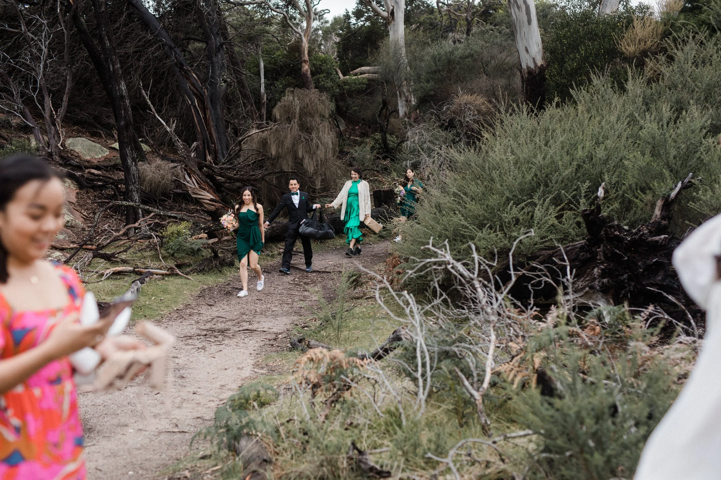 Group of people hiking on a trail through a wooded area, some dressed in formal attire, with trees and bushes surrounding them.
