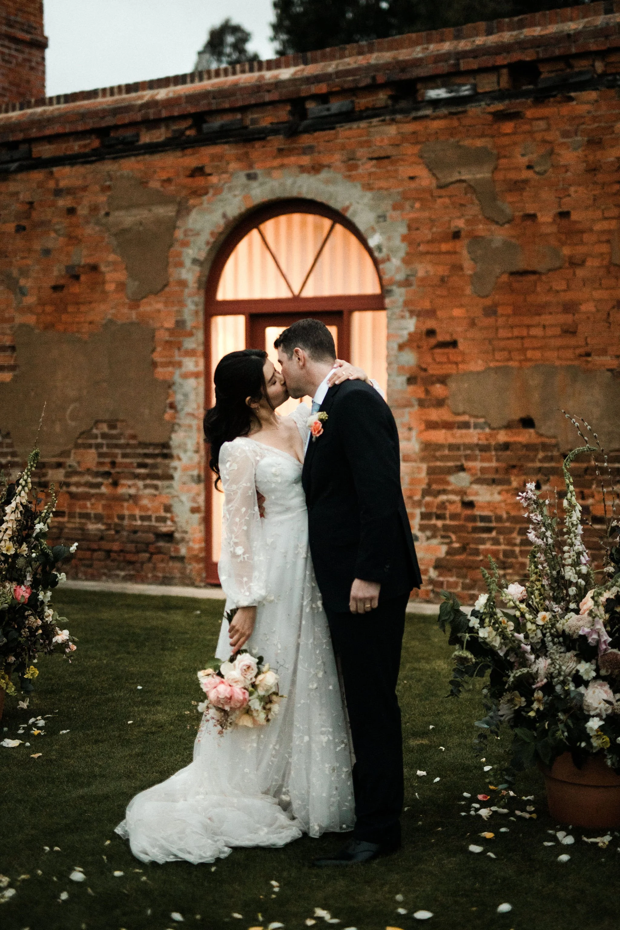 A bride and groom kiss outdoors in front of a rustic brick building during their wedding, surrounded by floral arrangements and flower petals on the grass.