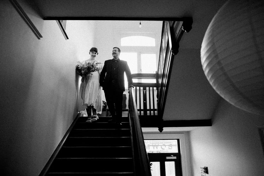 A woman in a white dress holding a bouquet and a man in a suit walking down a staircase inside a house