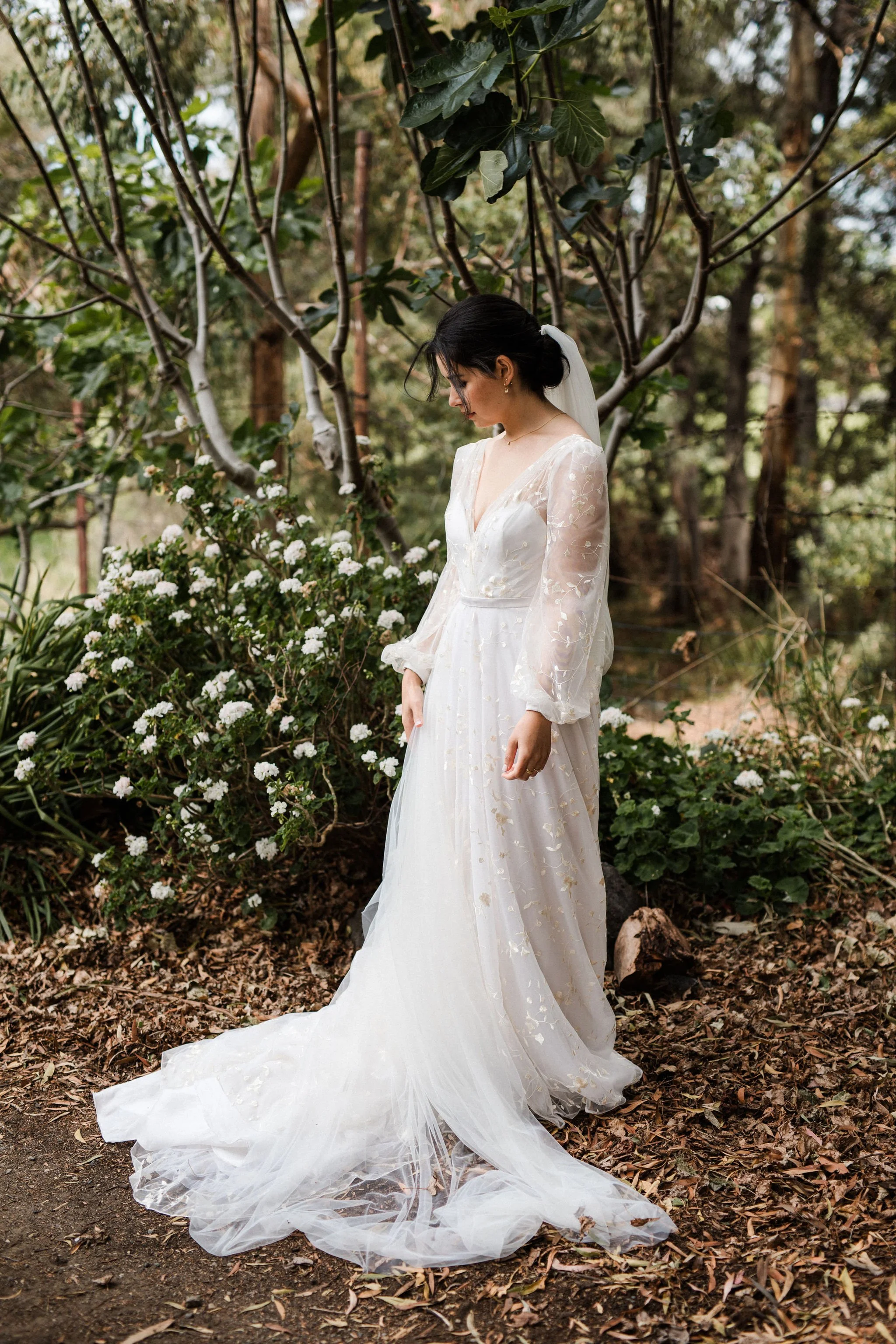 A woman in a white wedding dress standing in a forest, surrounded by greenery and white flowers, with a veil in her hair, looking down.