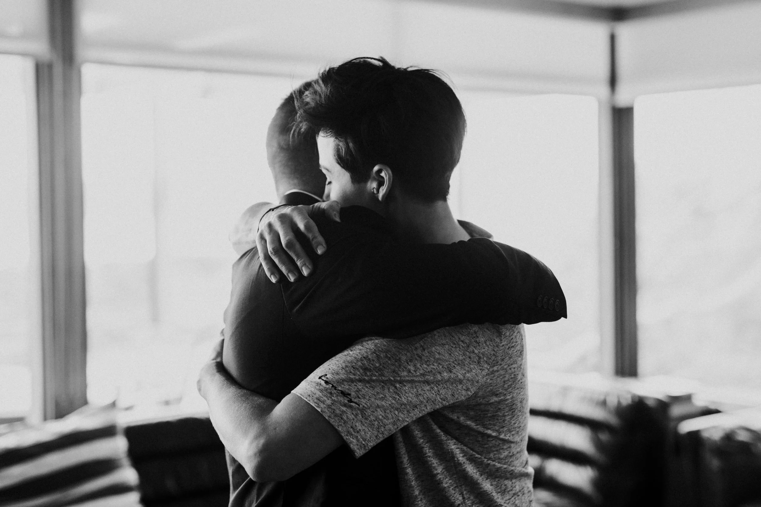 Two people hugging each other in a heartfelt embrace inside a room with large windows in the background, black and white photo.