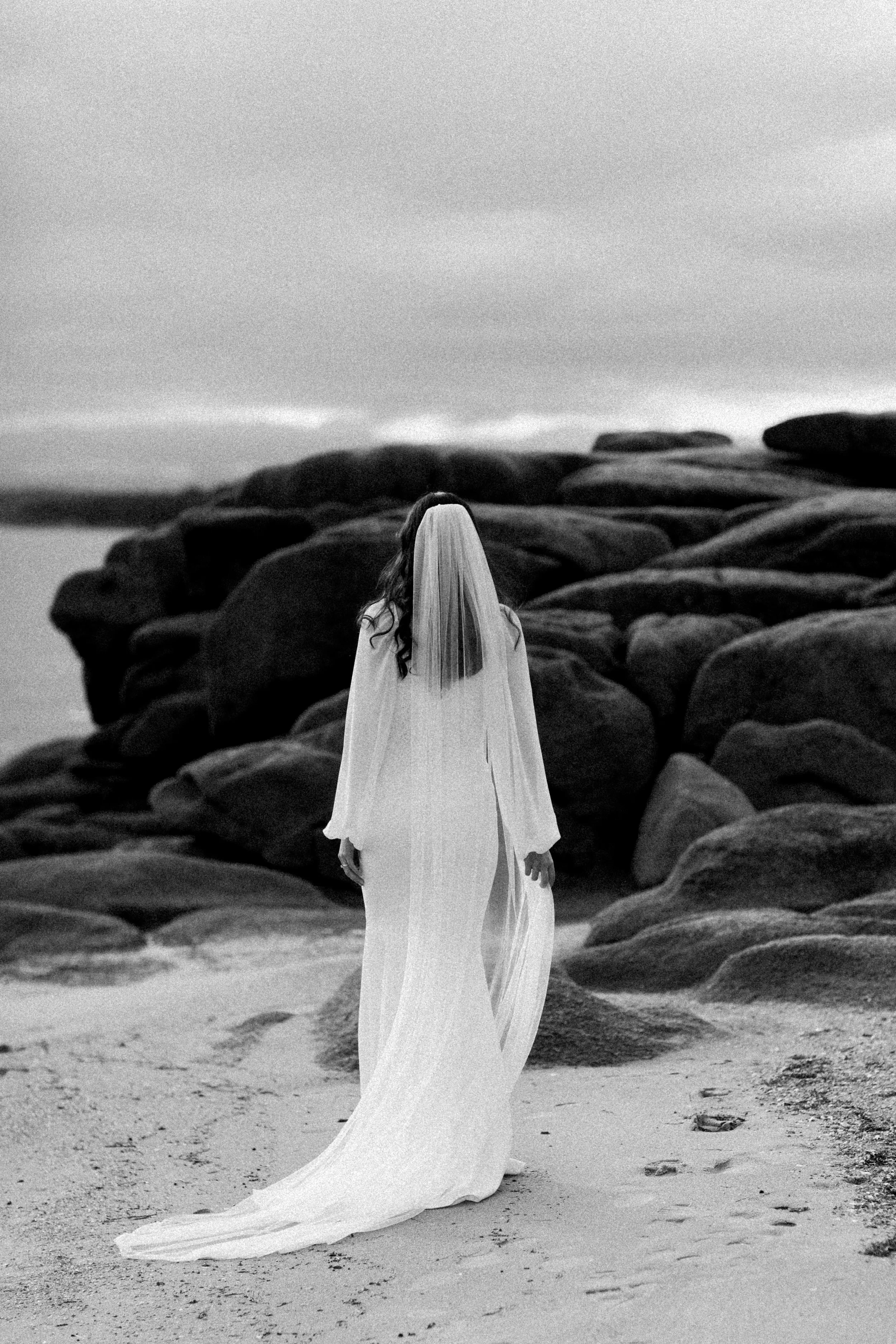 A woman in a wedding dress with a veil walking on a sandy beach with rocks and a cloudy sky in the background.
