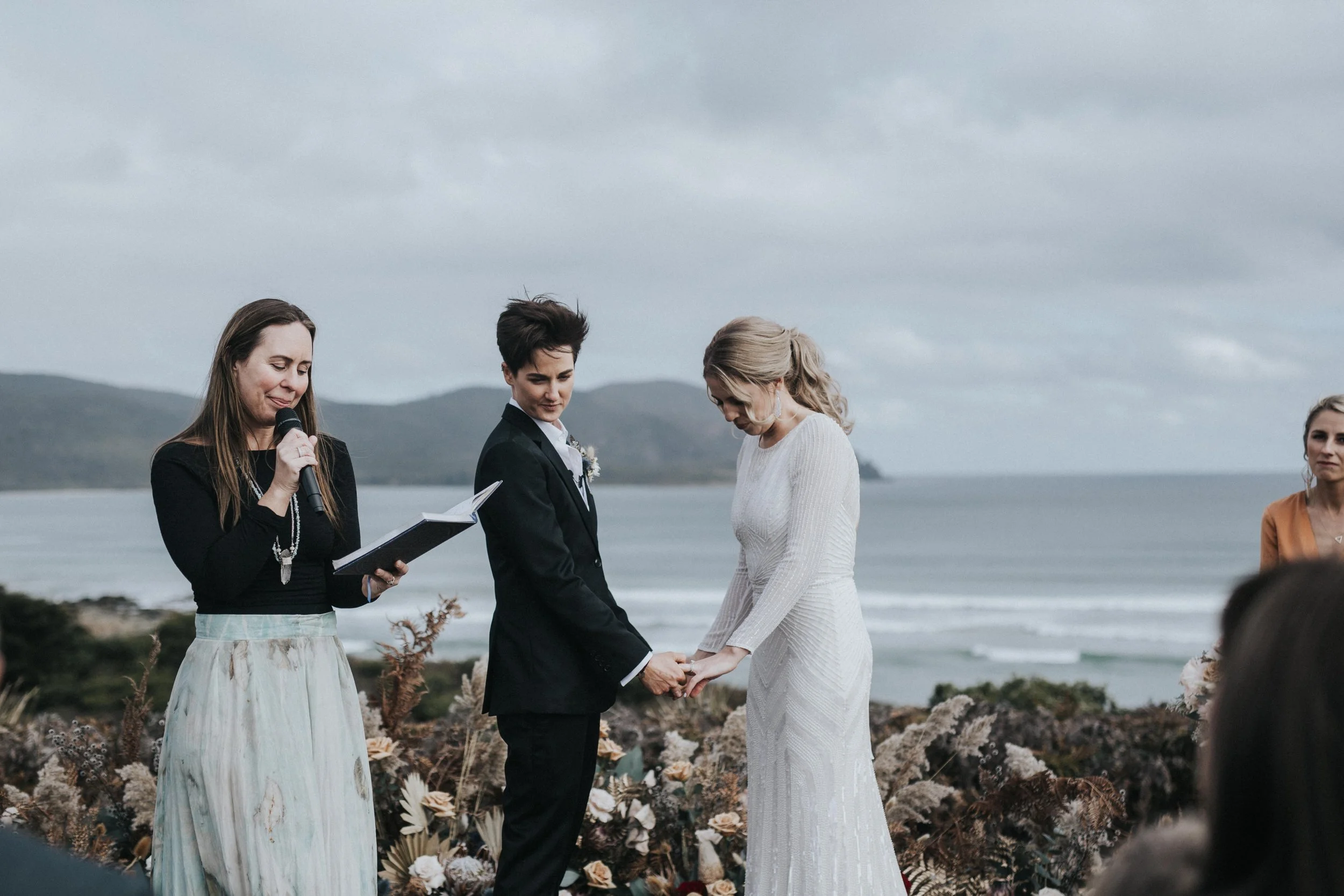A couple getting married outdoors near the ocean, holding hands, with an officiant reading vows and two women witnessing the ceremony.