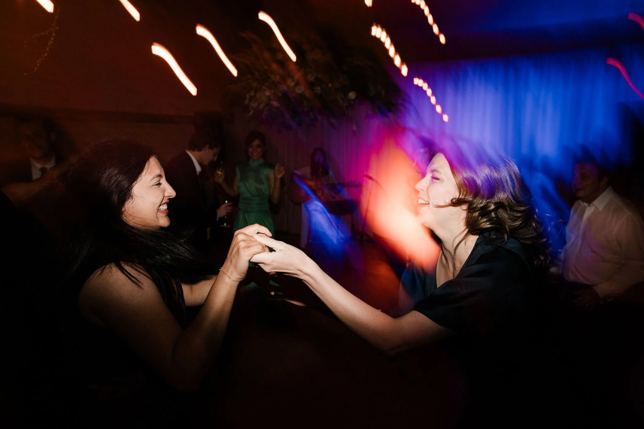 Two women dancing and holding hands at a party with colorful lights and other guests in the background.