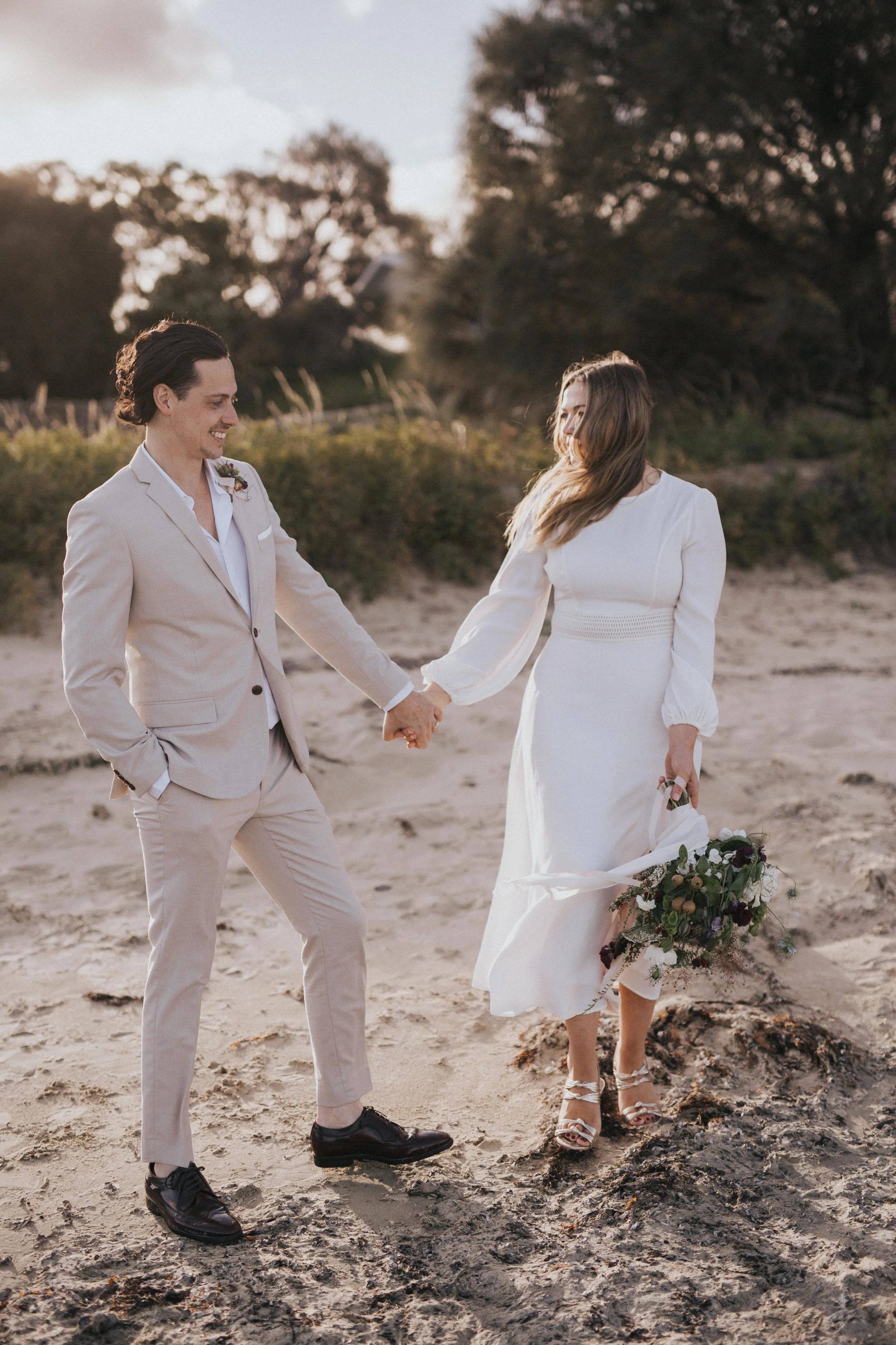 A bride and groom holding hands on a beach, smiling, during sunset. The bride is wearing a white dress and heels, holding a bouquet. The groom is in a light-colored suit and dark shoes.