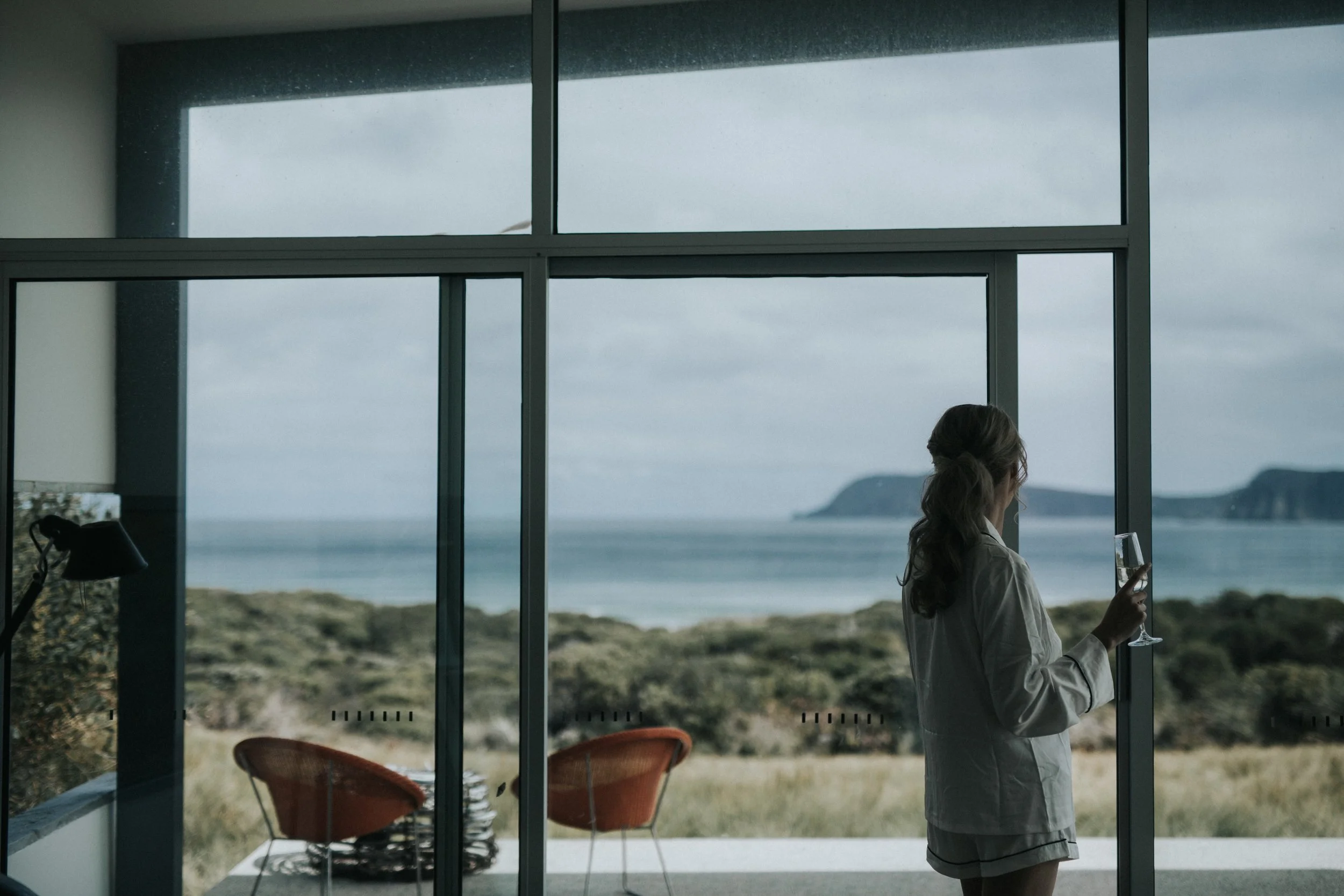 Woman with long hair in a white shirt holding a glass of water standing near large glass windows overlooking the ocean and distant cliffs