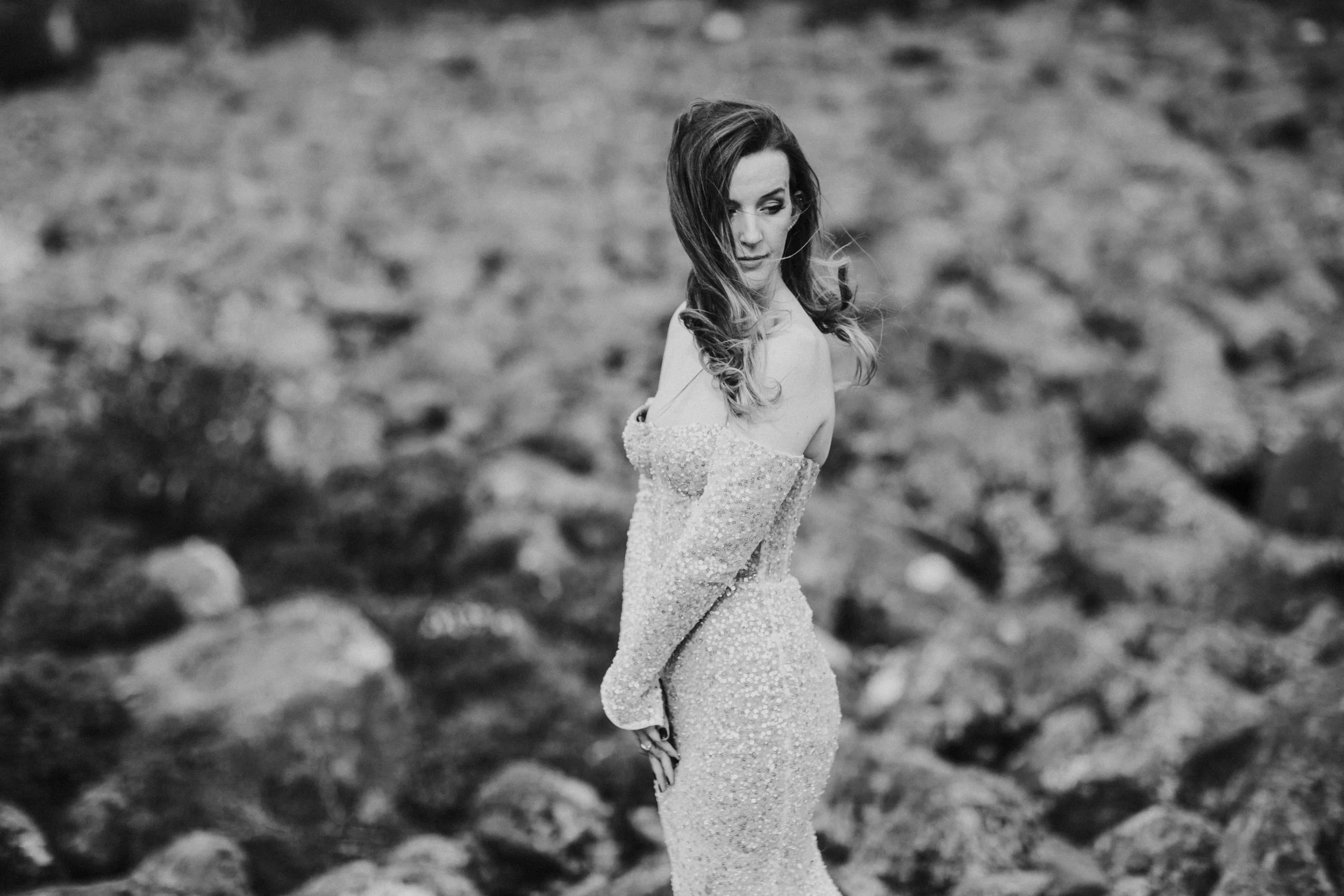 A black and white photograph of a woman standing outdoors on rocky terrain. She is wearing a long, off-shoulder, sparkling gown and has wavy hair. She is looking down with a contemplative expression.